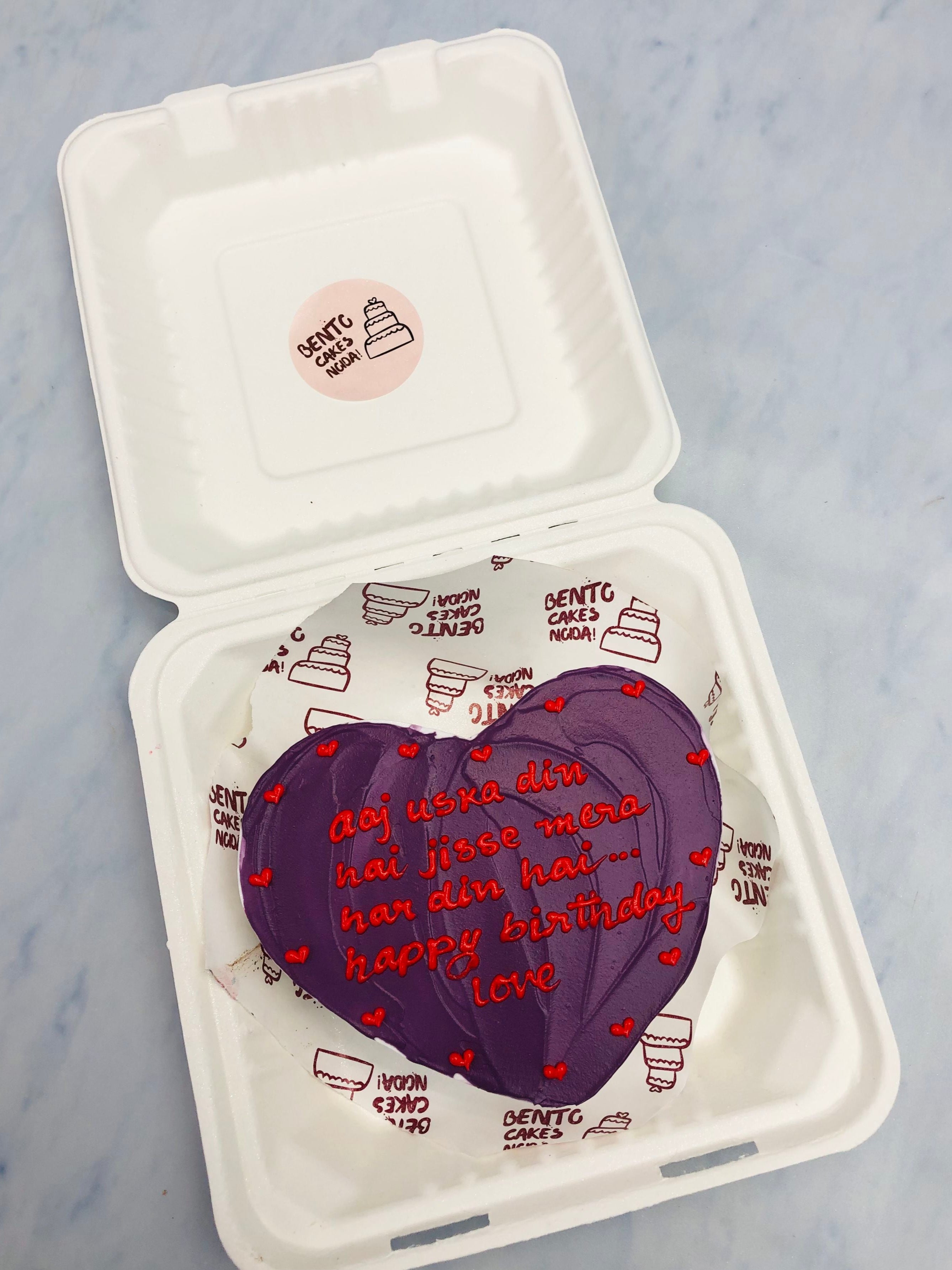 A dark purple heart shape cake with wavy design on top and filled with esthetic red hearts shape all over the cake and a loving text written with red icing.