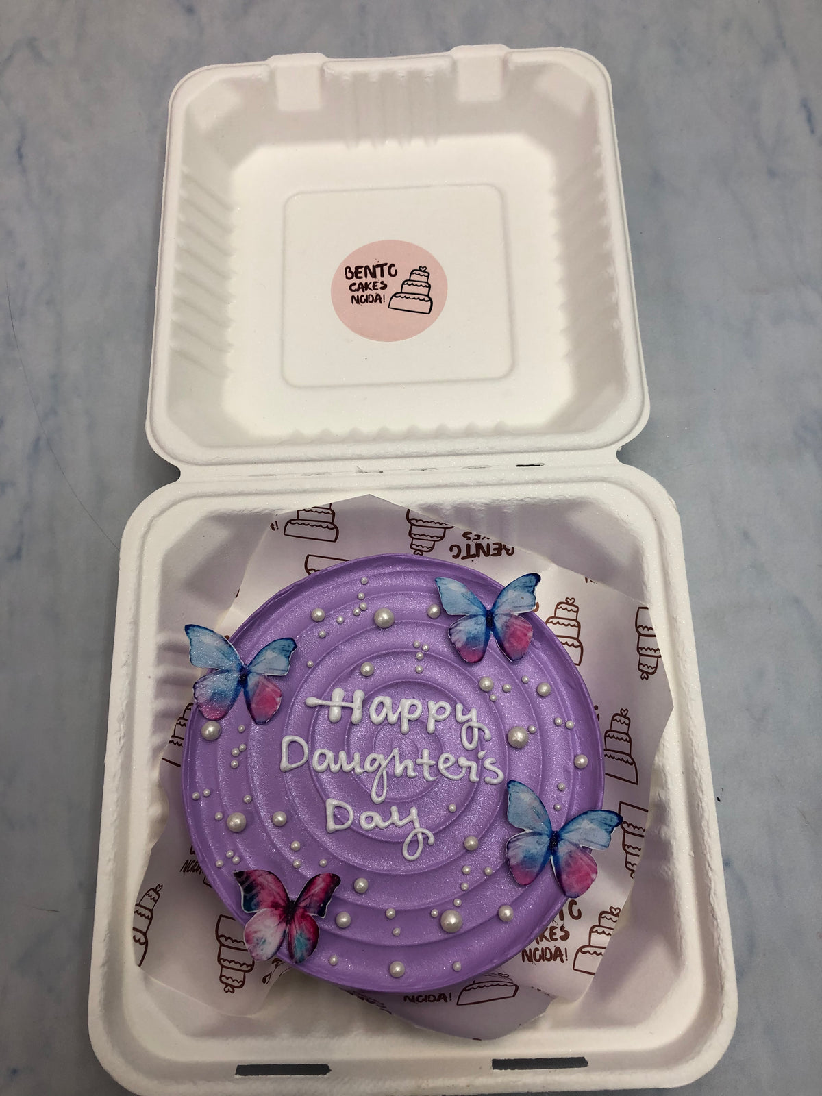 A shiney purple bento cake decorated with colorful butterflies and white sprinkles on it designed for Daughters day celebration.