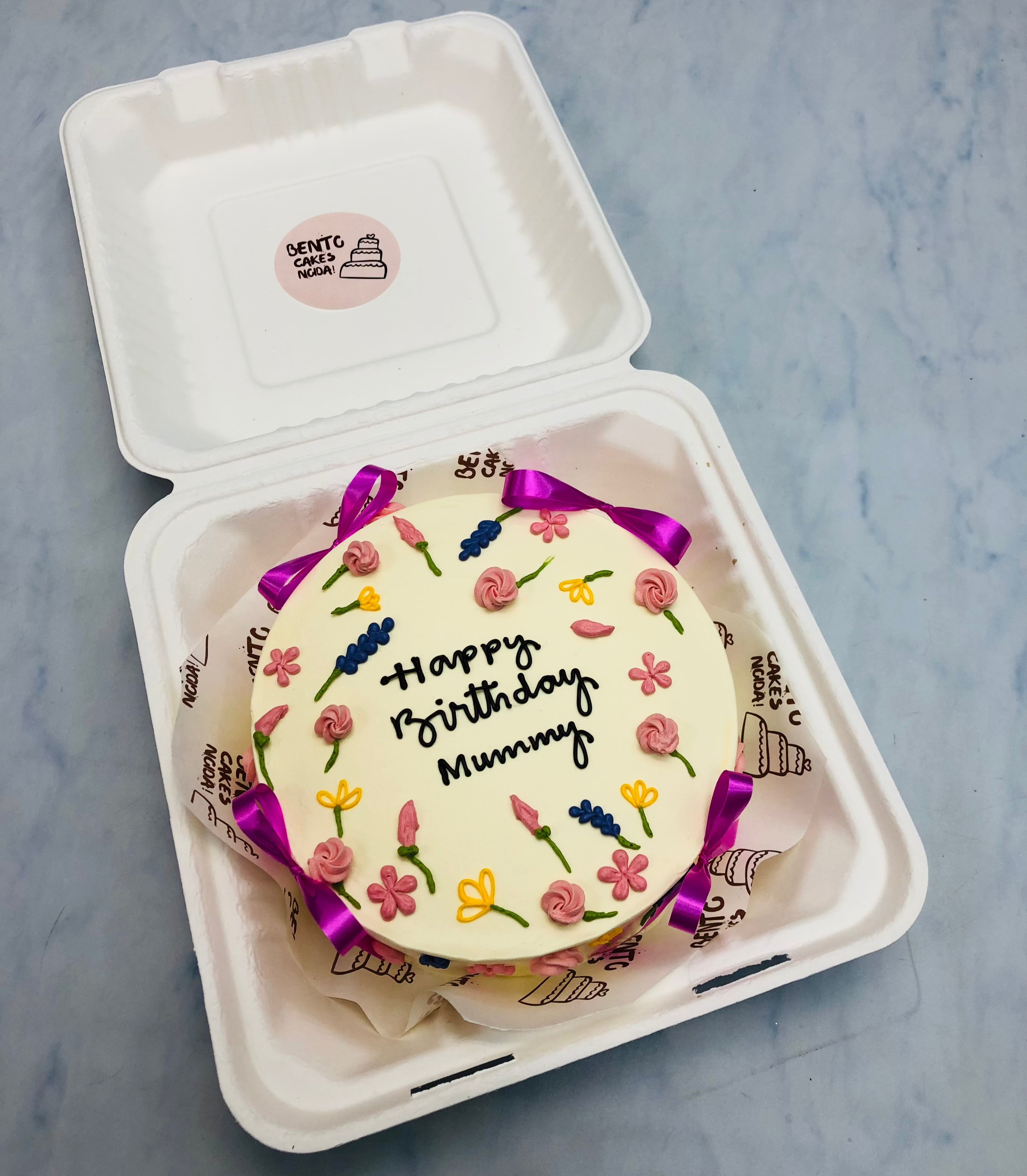 A beautifully decorated birthday cake with floral designs and the message "Happy Birthday Mummy" written on it, placed inside a bento box. The cake is adorned with pink, yellow, and blue flowers and purple ribbons.