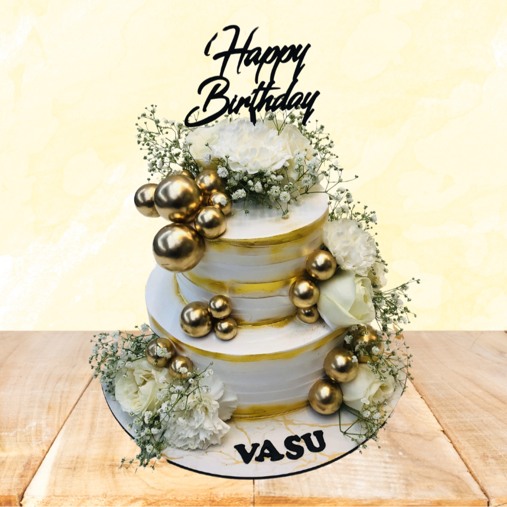 A three-tiered white and gold birthday cake adorned with white flowers, golden spherical decorations, and a "Happy Birthday" topper. The cake is labeled "VASU" and is marked as 100% eggless.