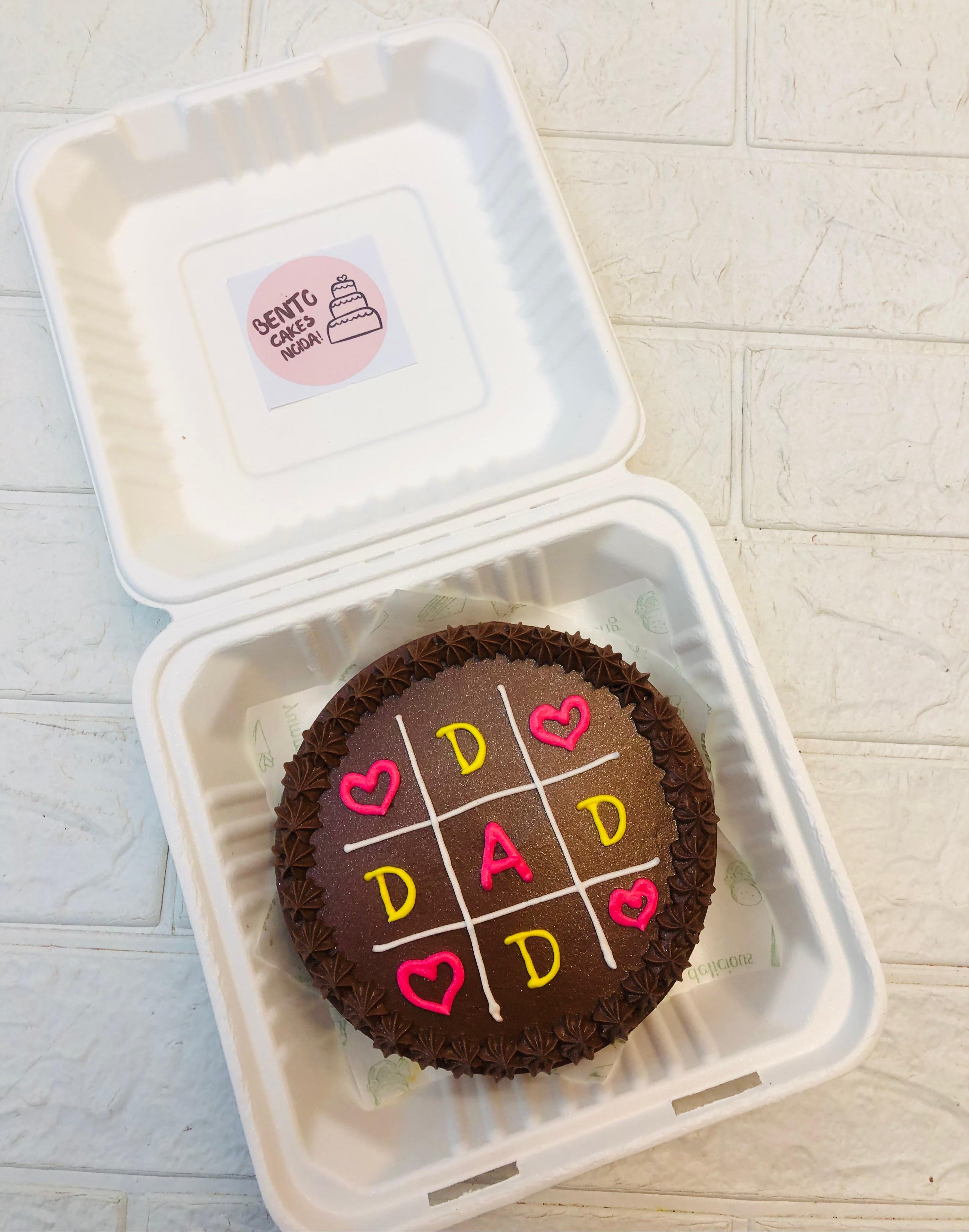 A chocolate bento cake decorated with a tic-tac-toe grid featuring the word "DAD" and heart symbols, placed inside a bento box.