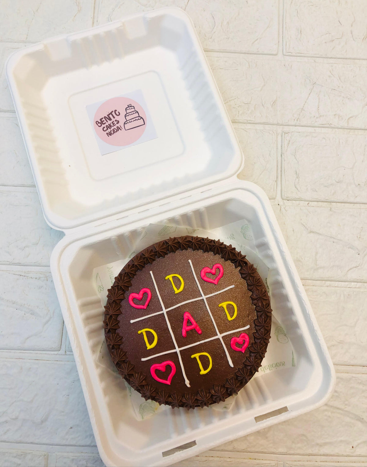 A chocolate bento cake decorated with a tic-tac-toe grid featuring the word "DAD" and heart symbols, placed inside a bento box.