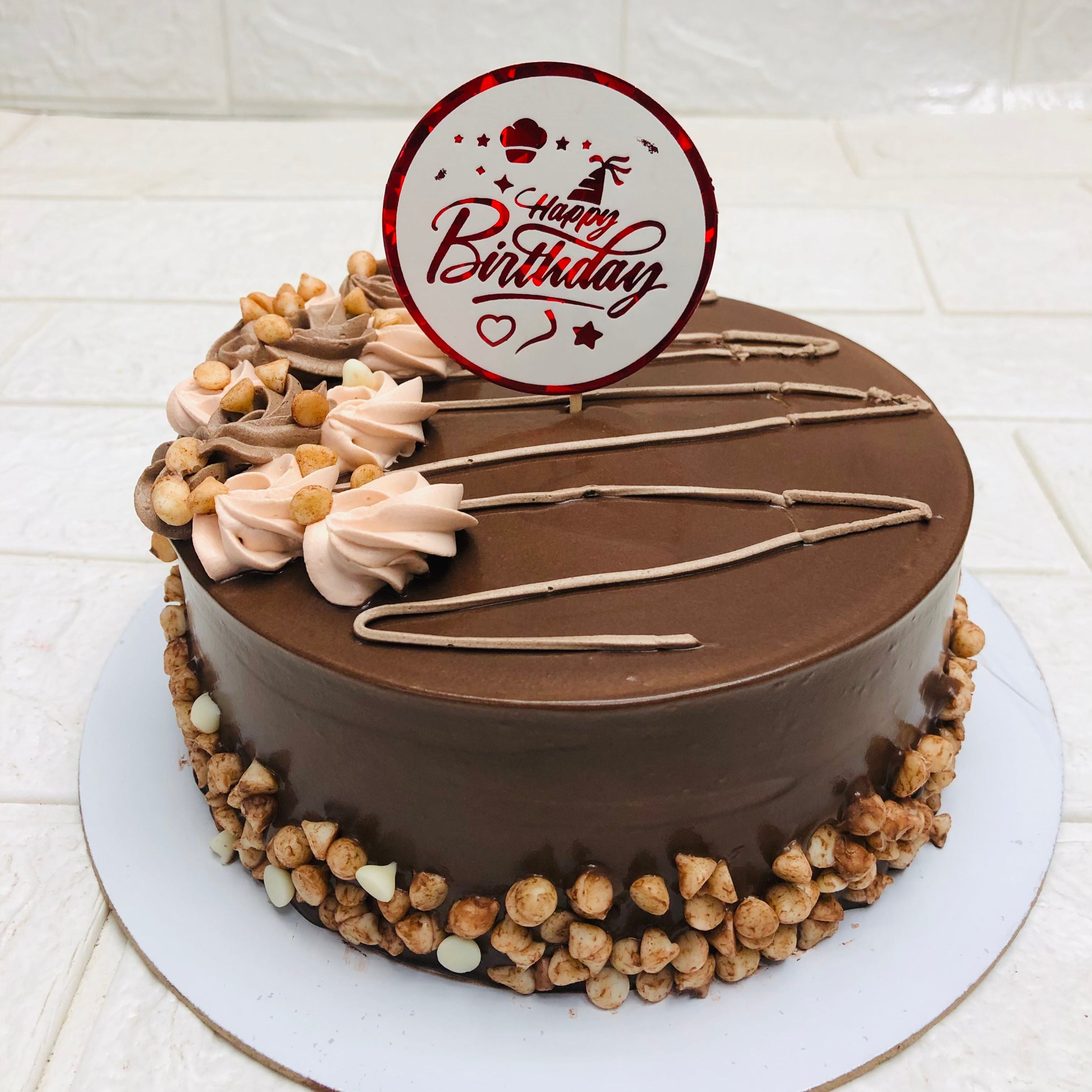 A beautifully decorated chocolate birthday cake with a glossy finish, topped with a "Happy Birthday" sign, swirls of light brown frosting, and surrounded by a border of chocolate chips and white chocolate drops.
