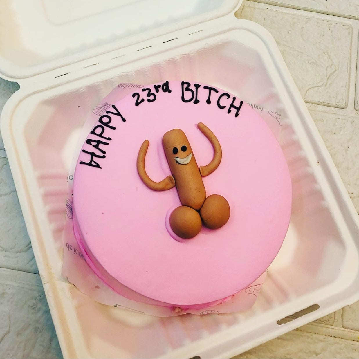 A pink cake with a humorous design featuring a smiling figure made of fondant, resembling male genitalia, and the text "HAPPY 23rd BITCH" written on top. The cake is placed inside a white takeout container.