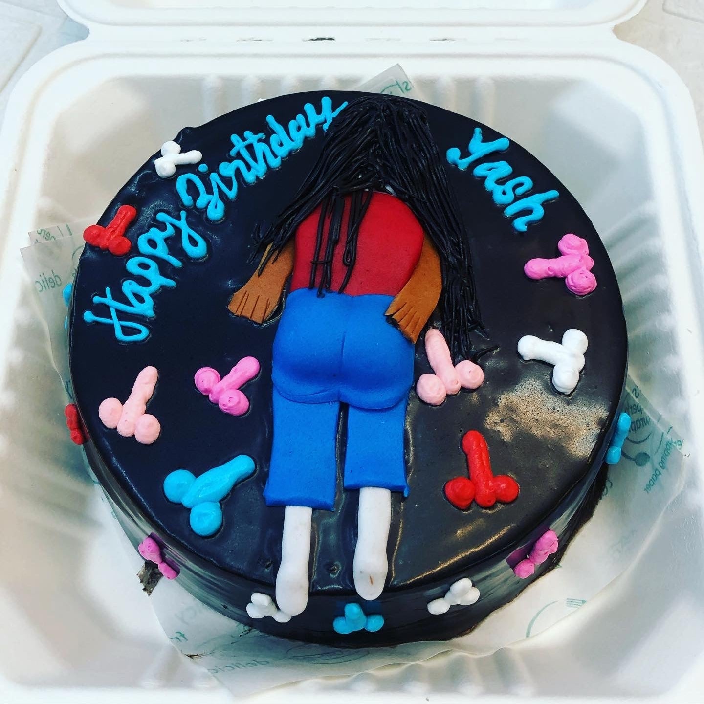 A chocolate cake with a fondant figure of a person lying face down, surrounded by colorful fondant shapes, inside a white box. The cake has the text "Happy Birthday Yash" written in blue icing.
