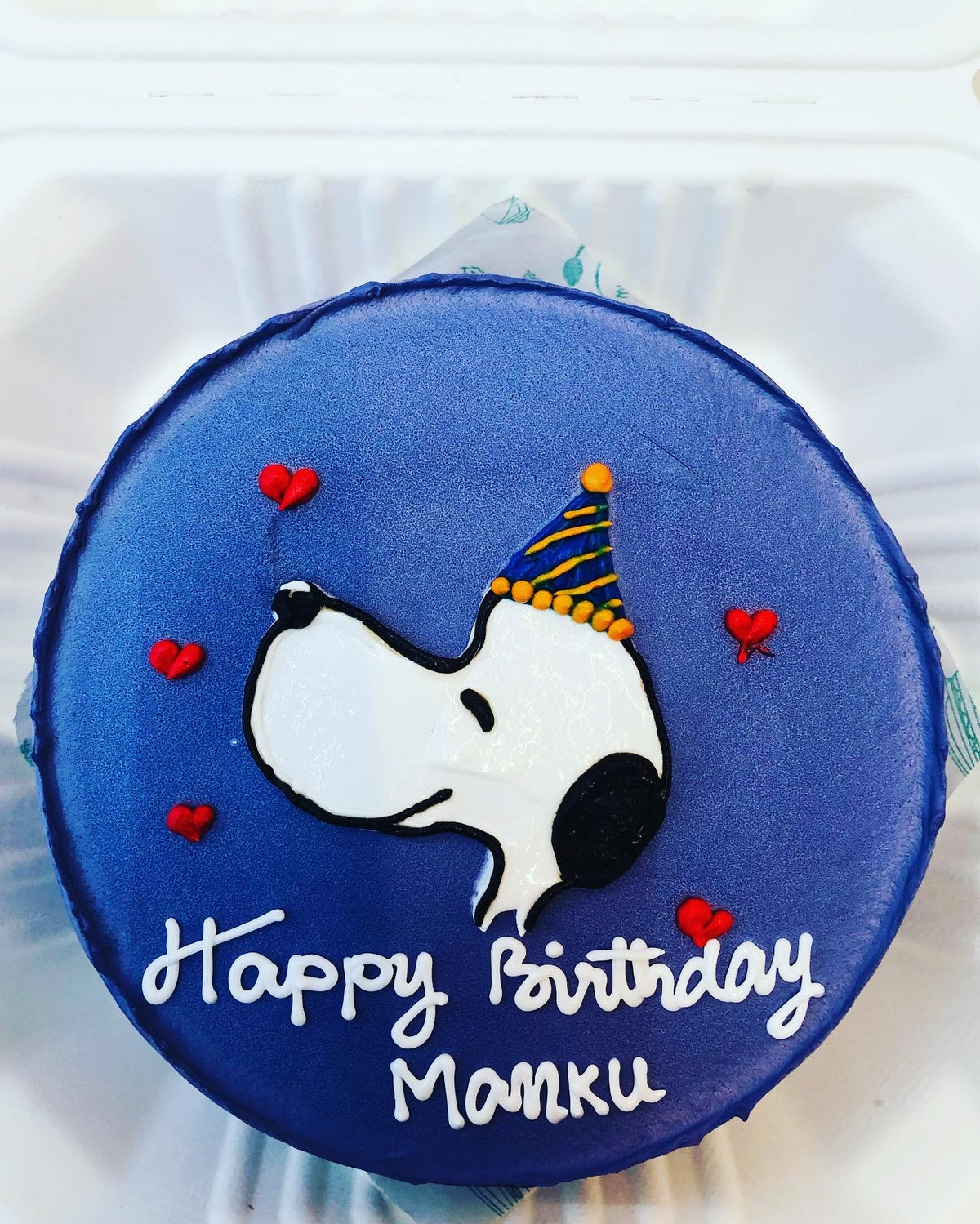 A round birthday cake with a blue frosting featuring a drawing of Snoopy wearing a party hat, surrounded by small red hearts, and the text "Happy Birthday Manku" written in white icing.
