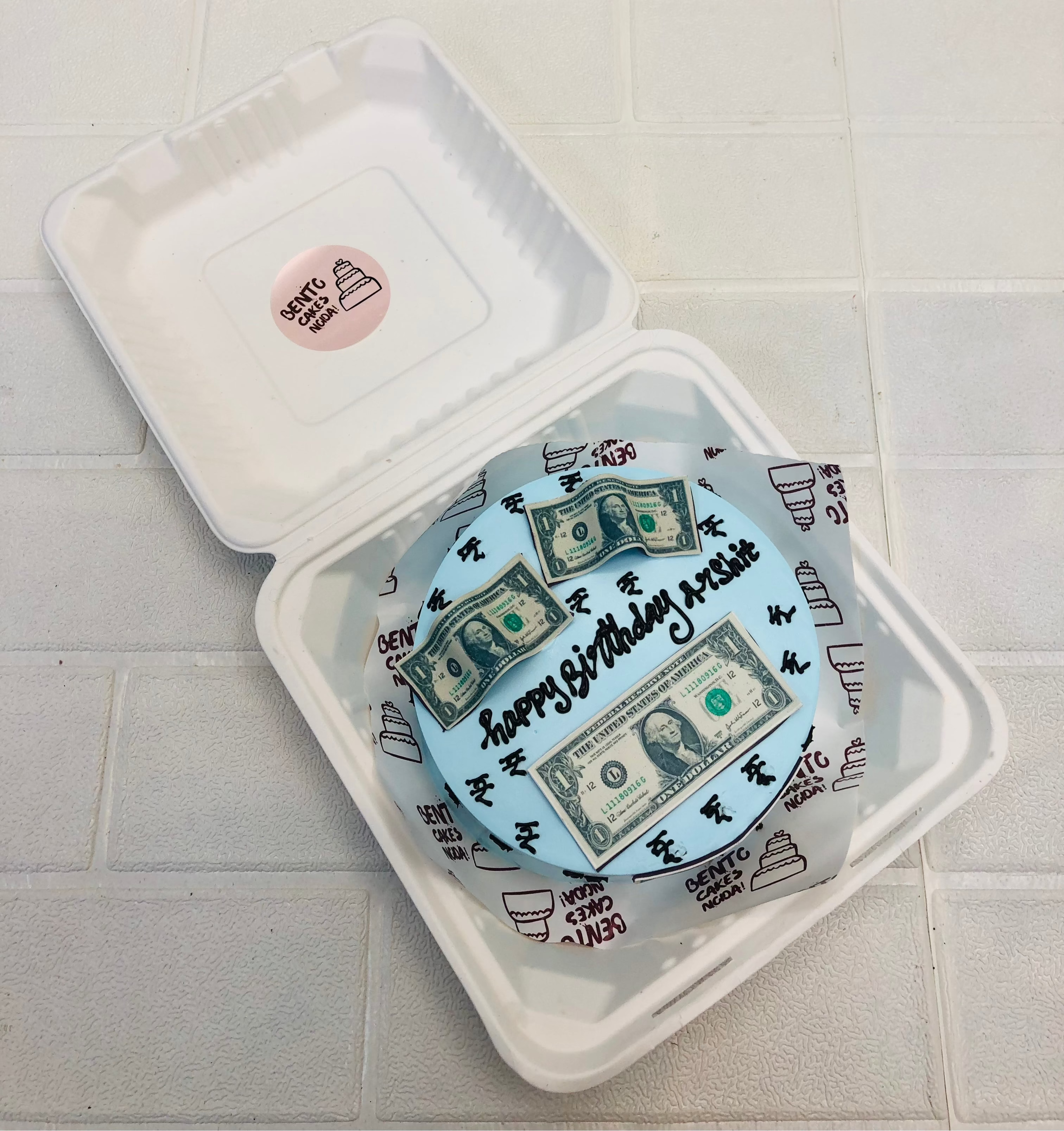 A round cake with light blue frosting, creatively decorated with realistic dollar bill images and the text "Happy Birthday Arshit" in black icing. 