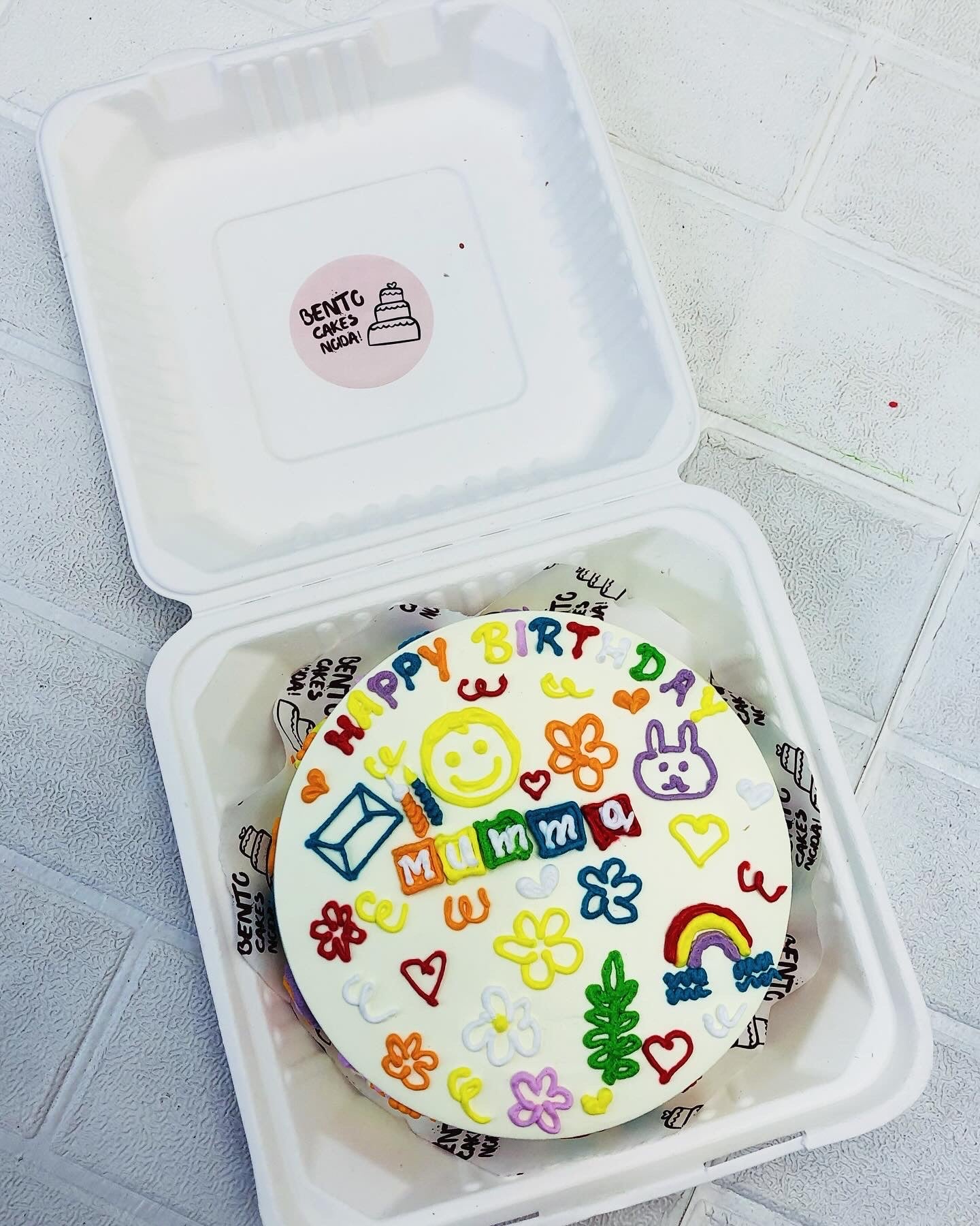 A cheerful birthday cake with vibrant icing designs, including flowers, hearts, a rainbow, a smiley face, a bunny, and an envelope, placed inside a white bento cake box. The cake features the text "Happy Birthday Mumma" and is set on a light-colored tiled surface.