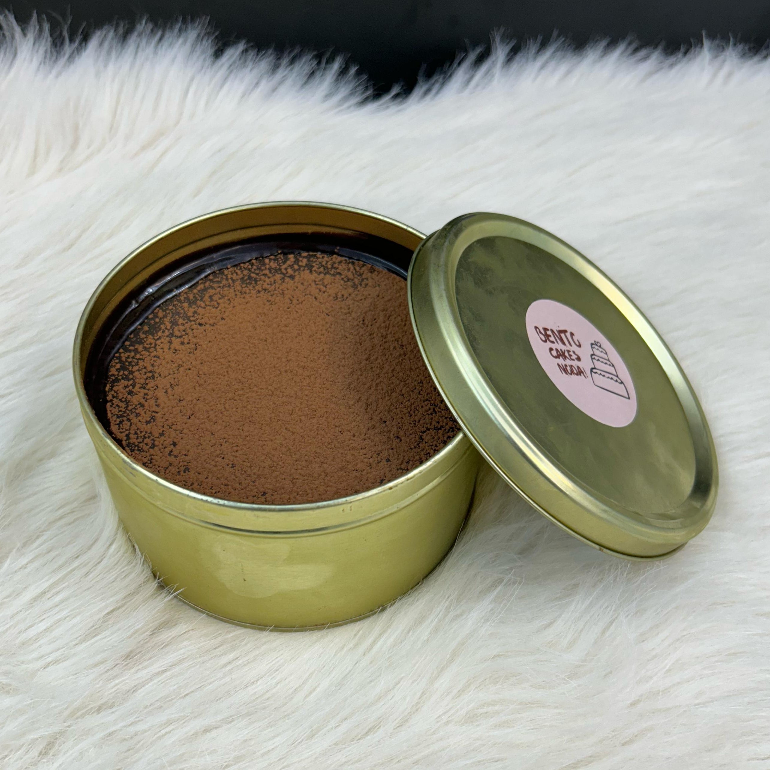 A luxurious Dream Tin Cake enclosed in an elegant golden tin, featuring a cracking chocolate disc, flowing chocolate liquid, luscious cream, and a soft, fluffy cake layer, displayed on a cozy white surface.