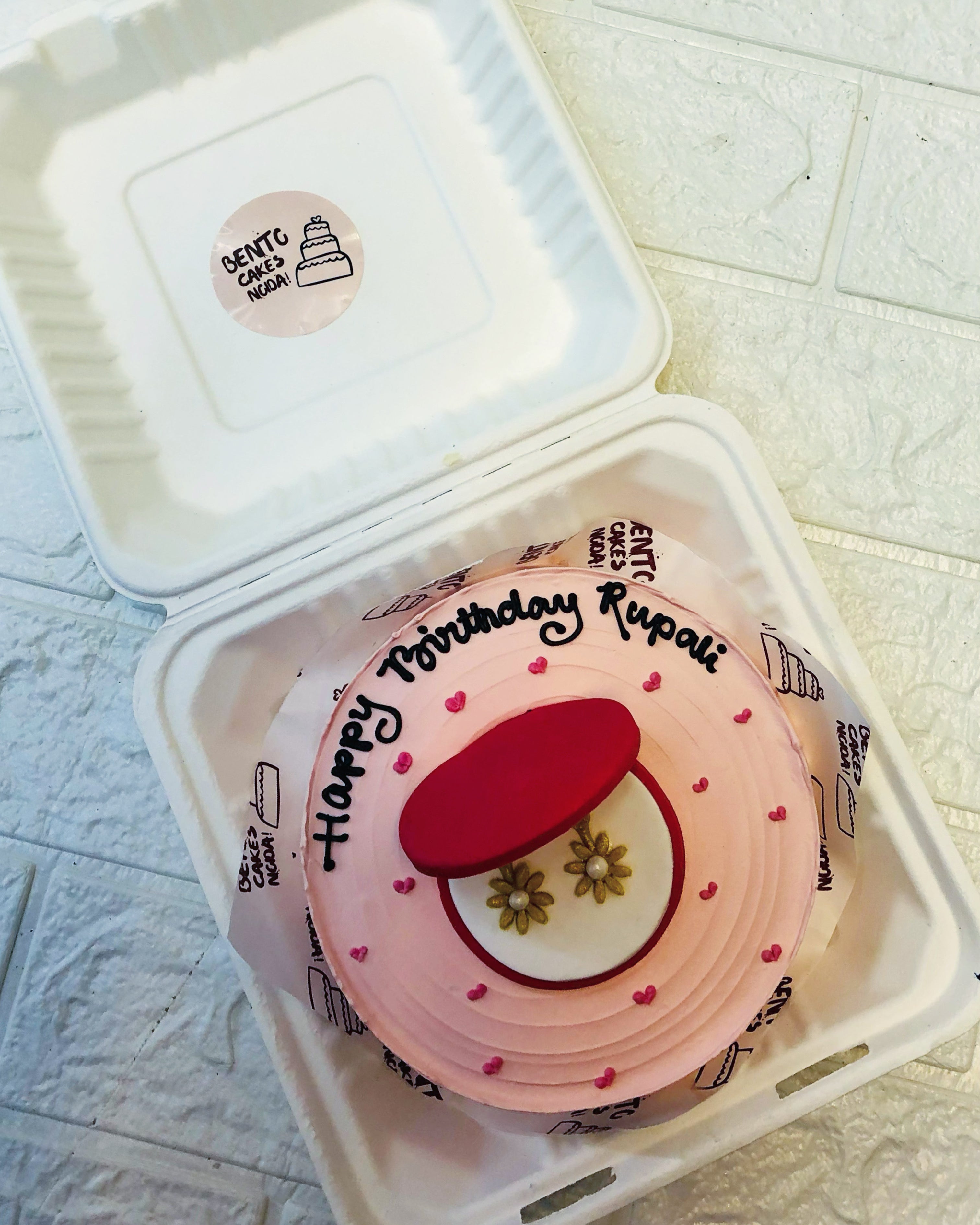 A pink bento cake with a birthday message for Rupali, placed inside a white bento box. The cake is decorated with a red circular topper and two flower designs. 
