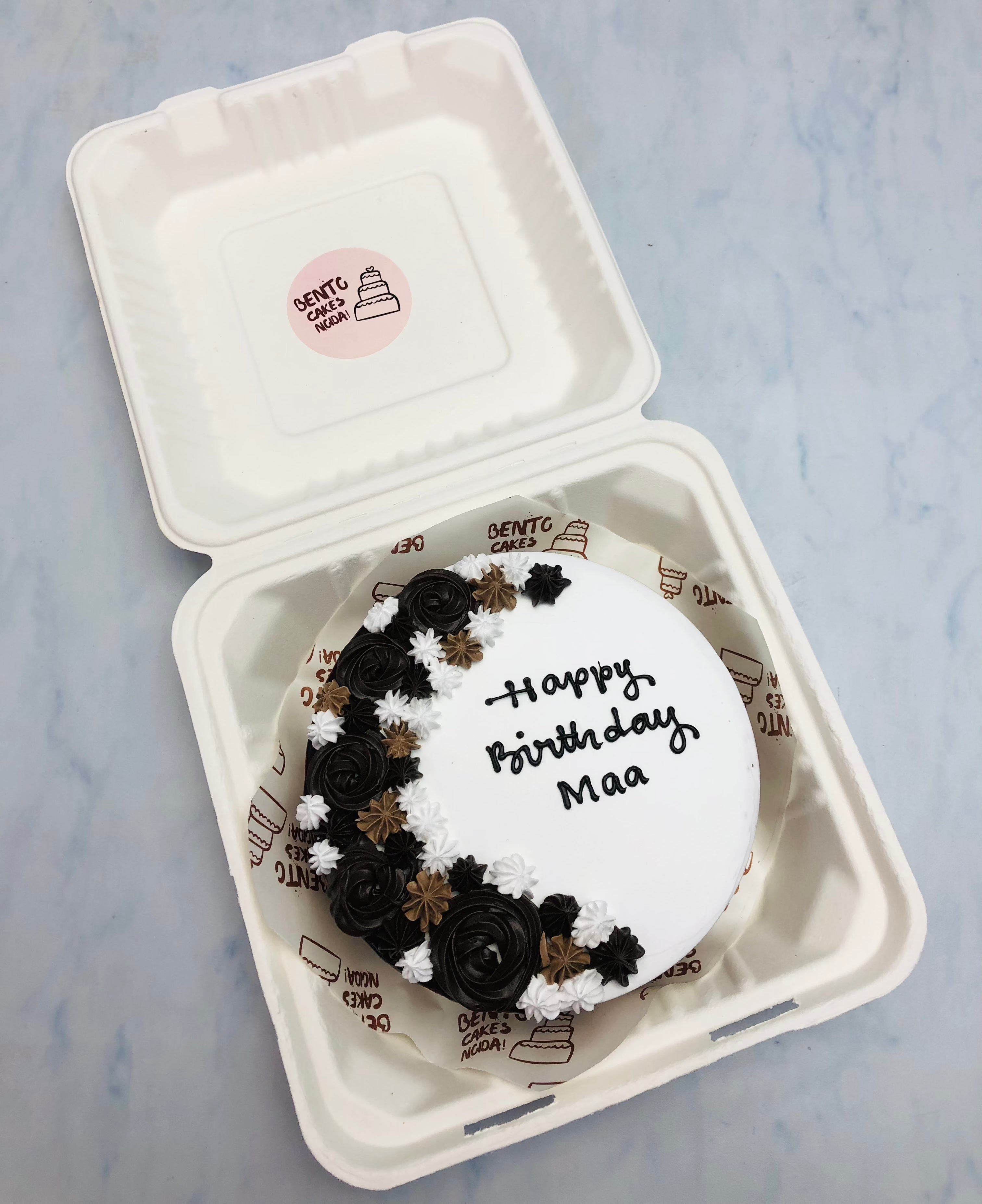 A beautifully crafted white bento cake with delicate floral decorations in black, brown, and white. The cake features the message "Happy Birthday Maa".