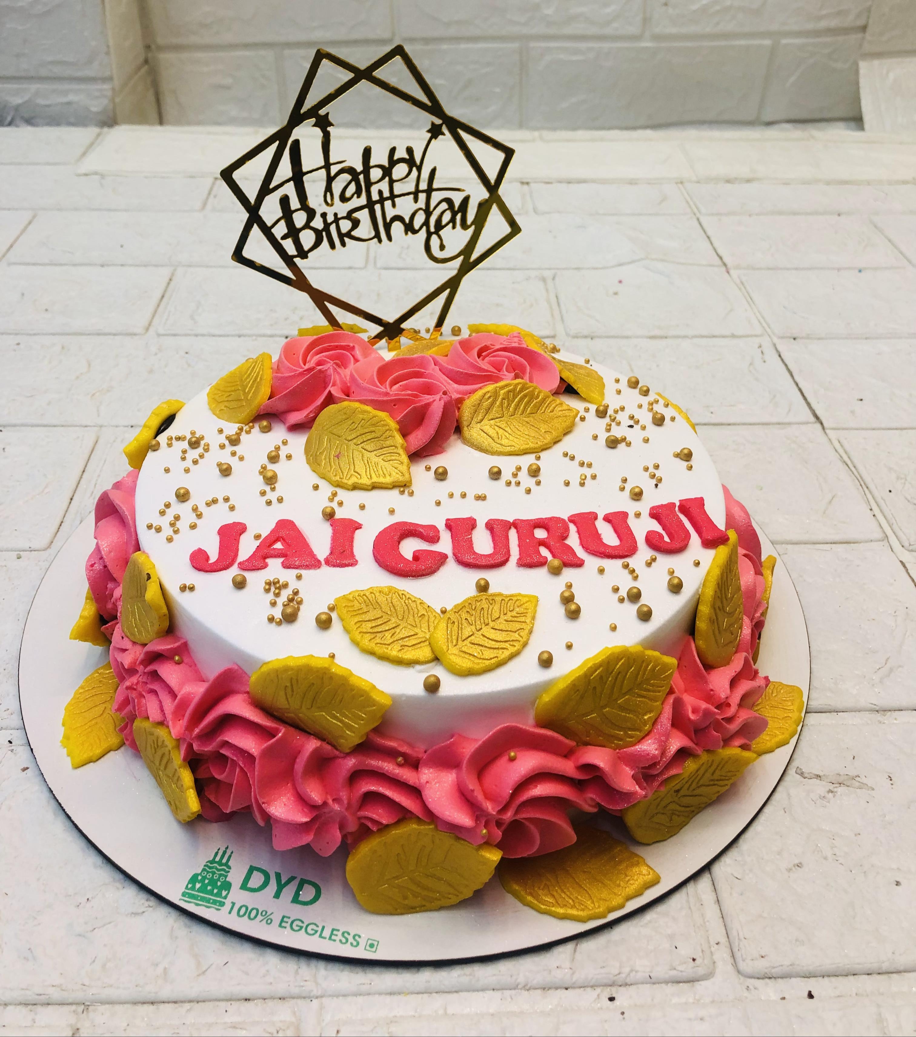 A stunning birthday cake featuring the message 'JAI GURUJI' in red icing. The cake is adorned with pink frosting swirls, gold decorative leaves, and small gold beads. A gold geometric topper with 'Happy Birthday' sits gracefully on top.