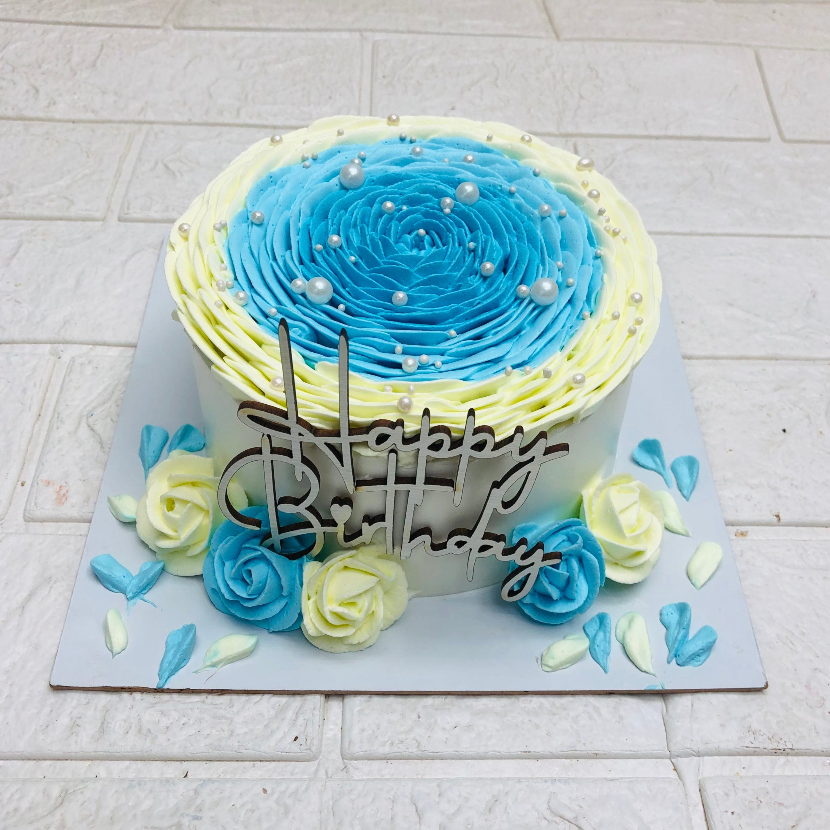 A beautifully decorated birthday cake with a "Happy Birthday" topper. The cake features a gradient of blue and yellow frosting with pearl-like decorations and is surrounded by blue and yellow buttercream roses and petals.