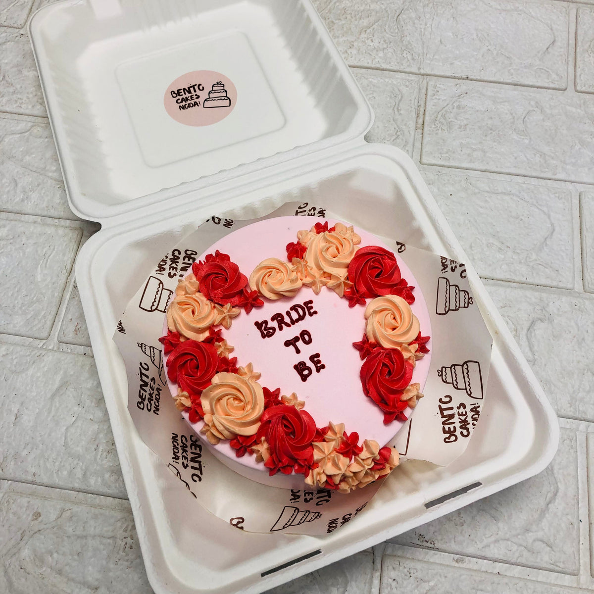 A beautifully decorated bento cake with the words "BRIDE TO BE" written on it, surrounded by red and peach-colored floral icing designs, placed in a white takeout box.