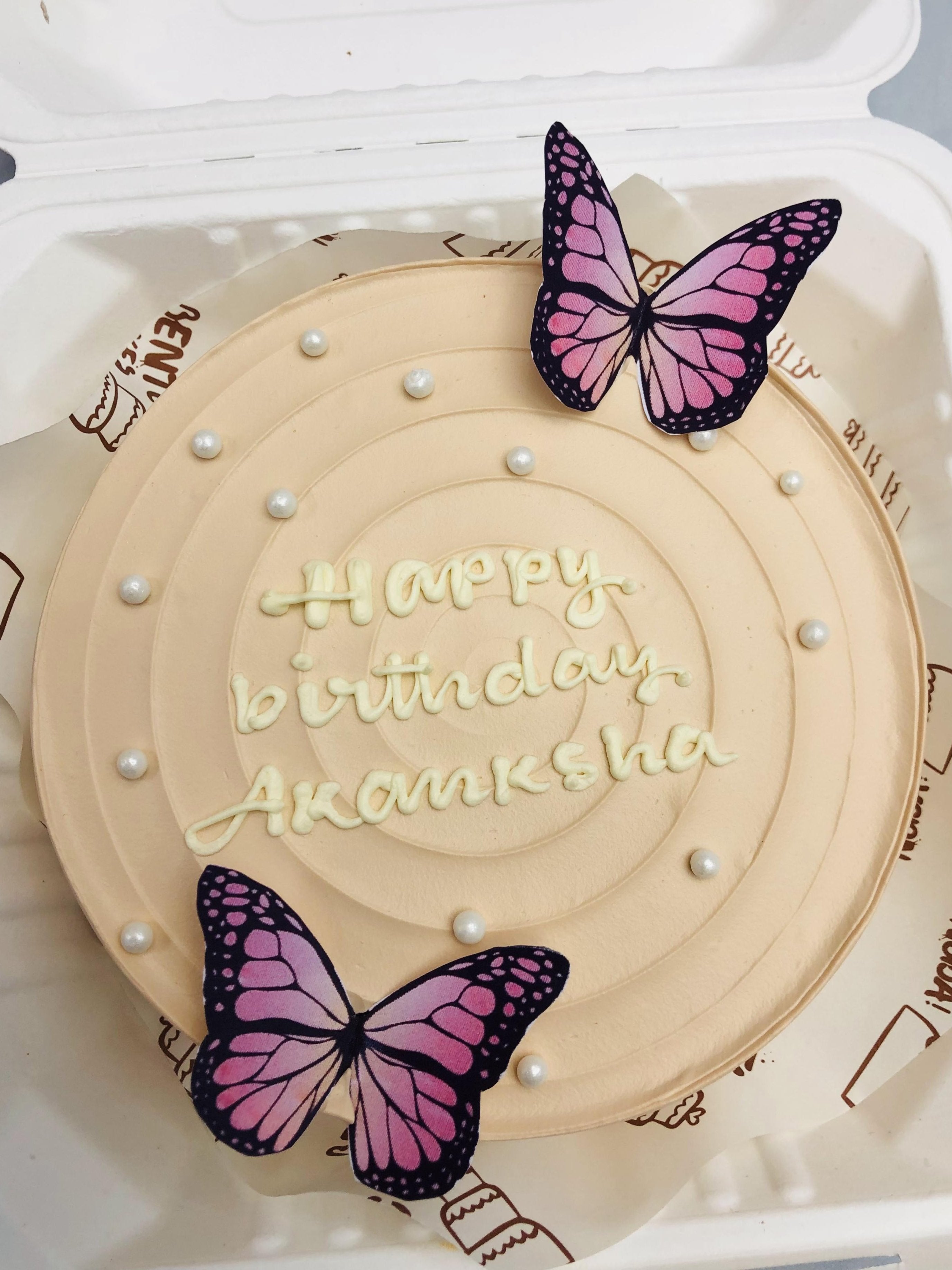 A beautifully decorated birthday cake with the message "Happy birthday Akanksha" written in white icing. The cake is adorned with two realistic-looking pink and purple butterfly decorations and small white pearl-like embellishments.