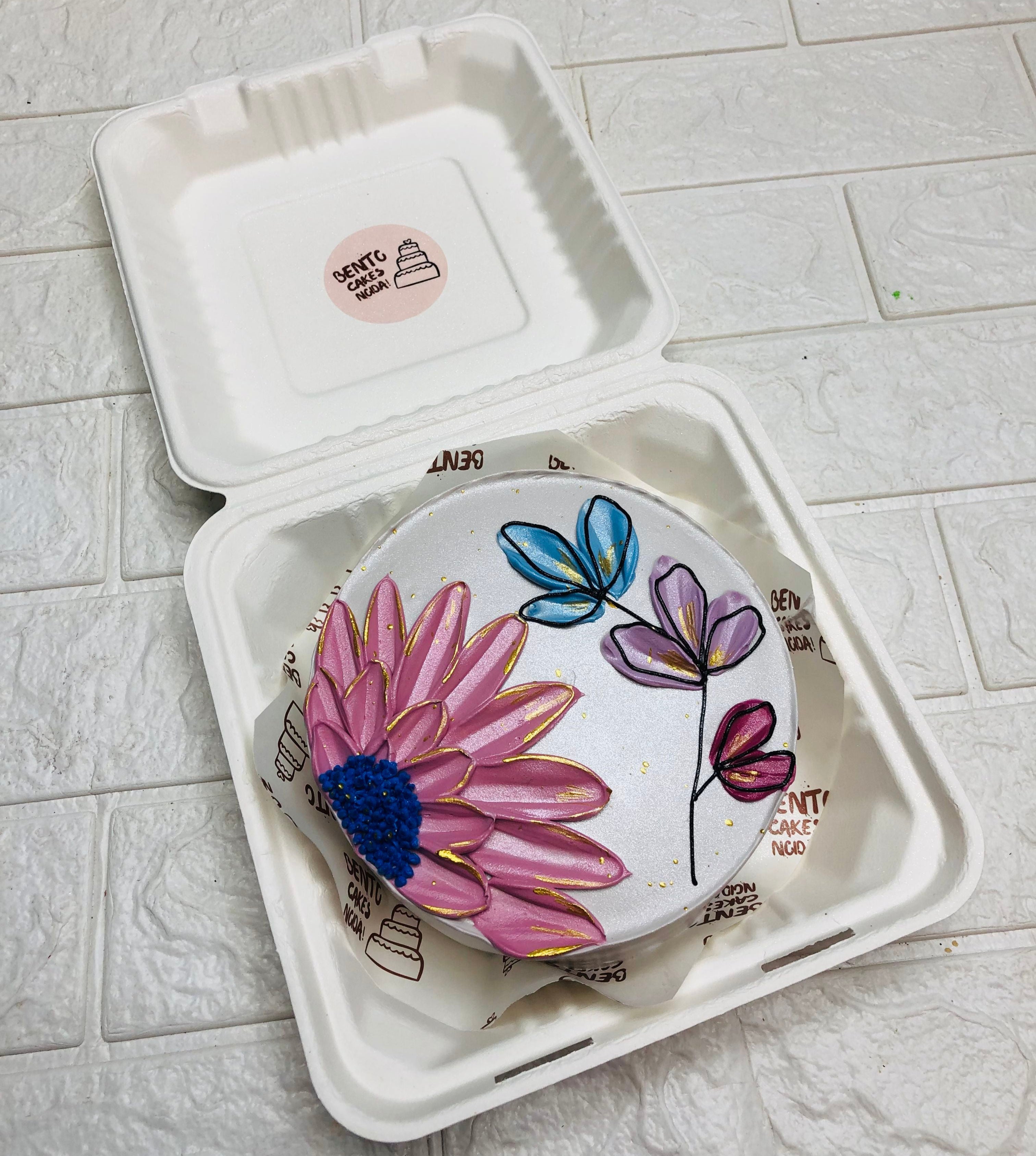 A beautifully decorated bento cake inside a white biodegradable container. The cake features intricate floral designs with a large pink flower at the center, surrounded by smaller blue, purple, and pink blossoms.