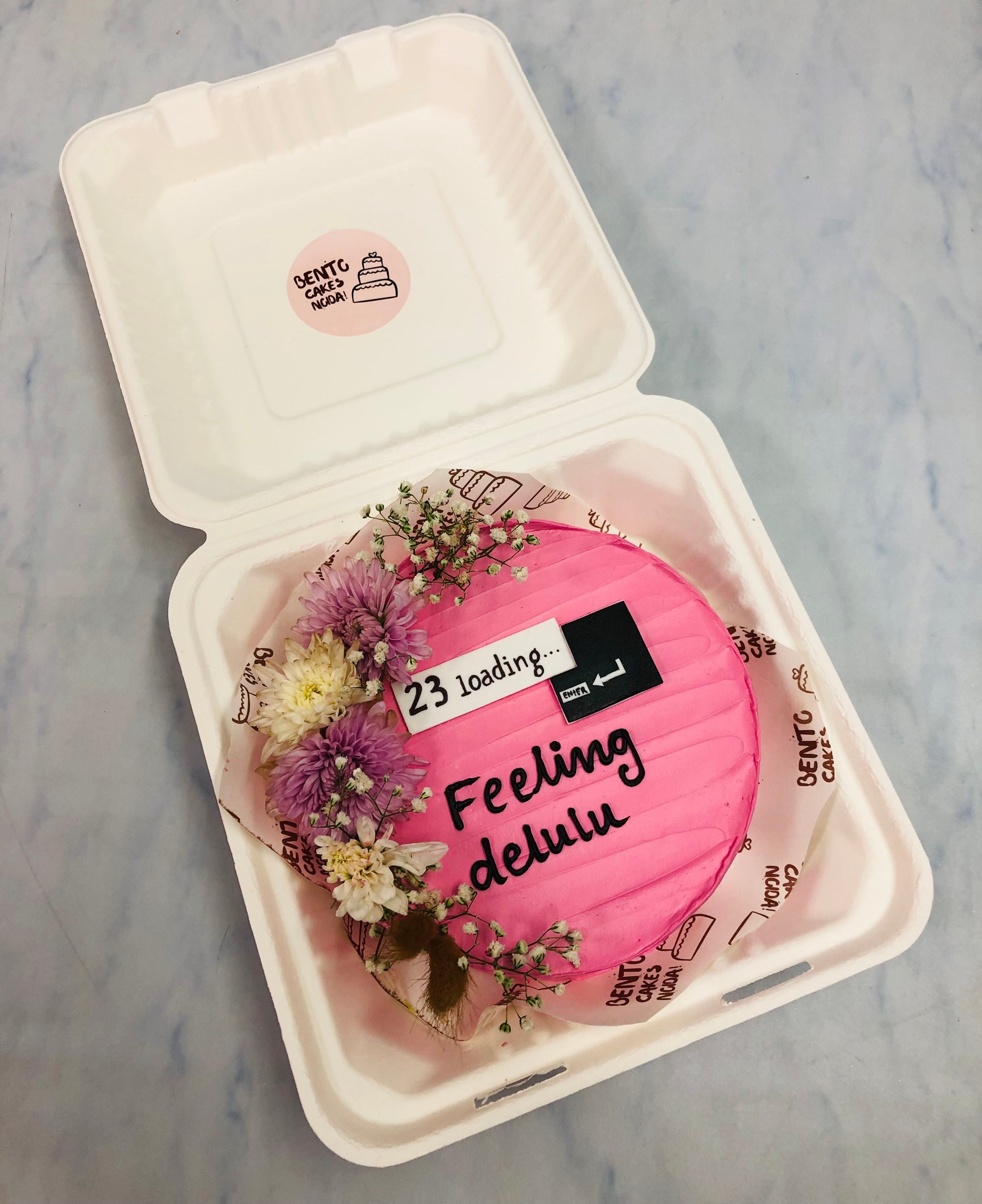 A round pink cake adorned with delicate floral decorations inside a white box. The cake features the humorous text '23 loading... Feeling delulu' alongside a progress bar graphic.
