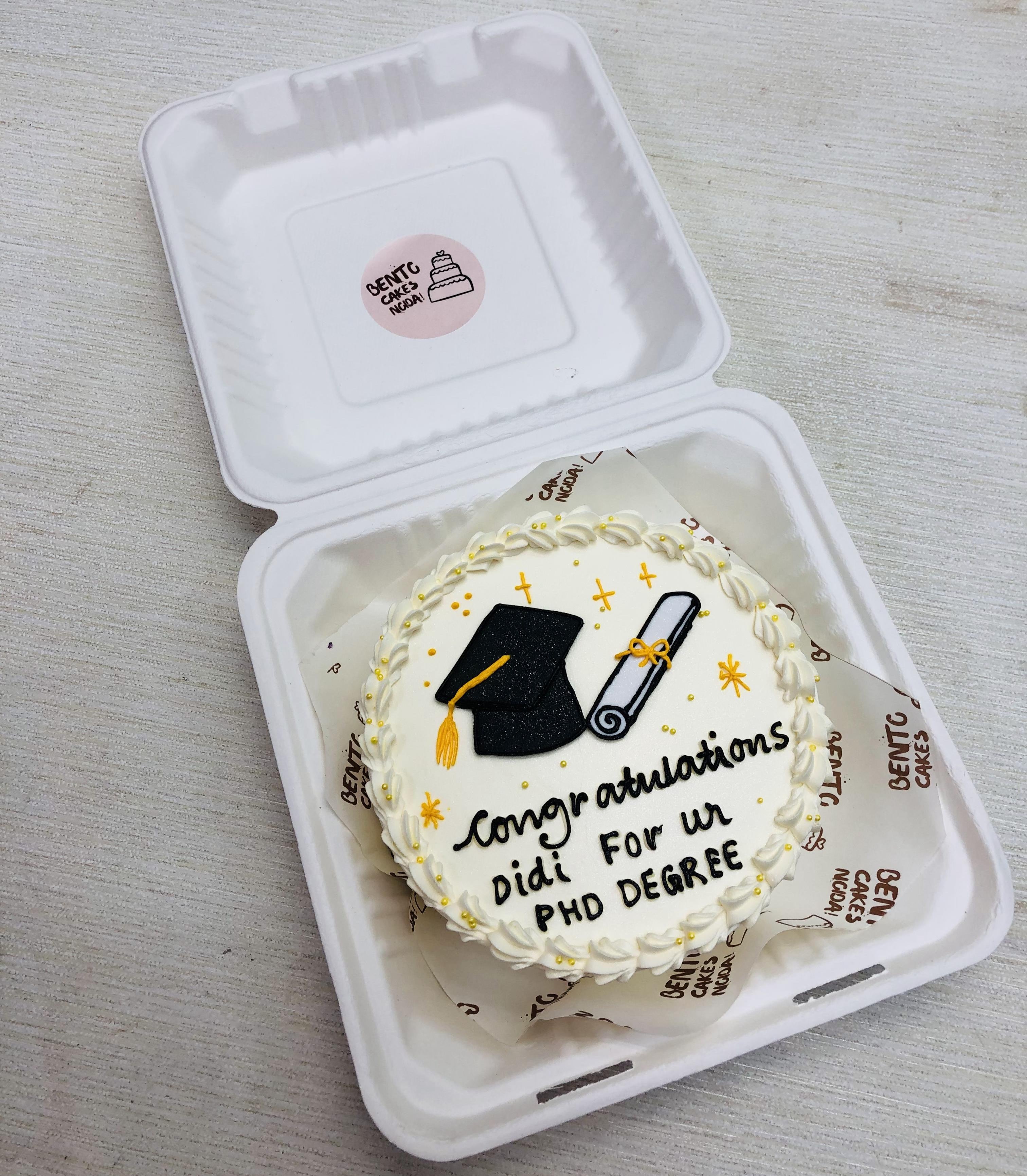 A beautifully crafted round cake with white icing, celebrating a PhD achievement. The cake is adorned with a black graduation cap, a rolled diploma, and yellow stars.