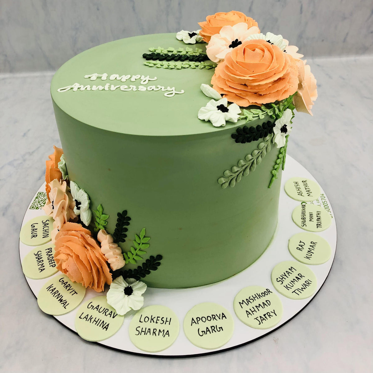 A beautifully designed green anniversary cake adorned with peach and white floral decorations. The top of the cake features the message "Happy Anniversary" in white icing, while the base is surrounded by circular plaques, each displaying a personalized name. This artistic cake combines elegance with a personal touch, making it a perfect centerpiece for celebrating special occasions.