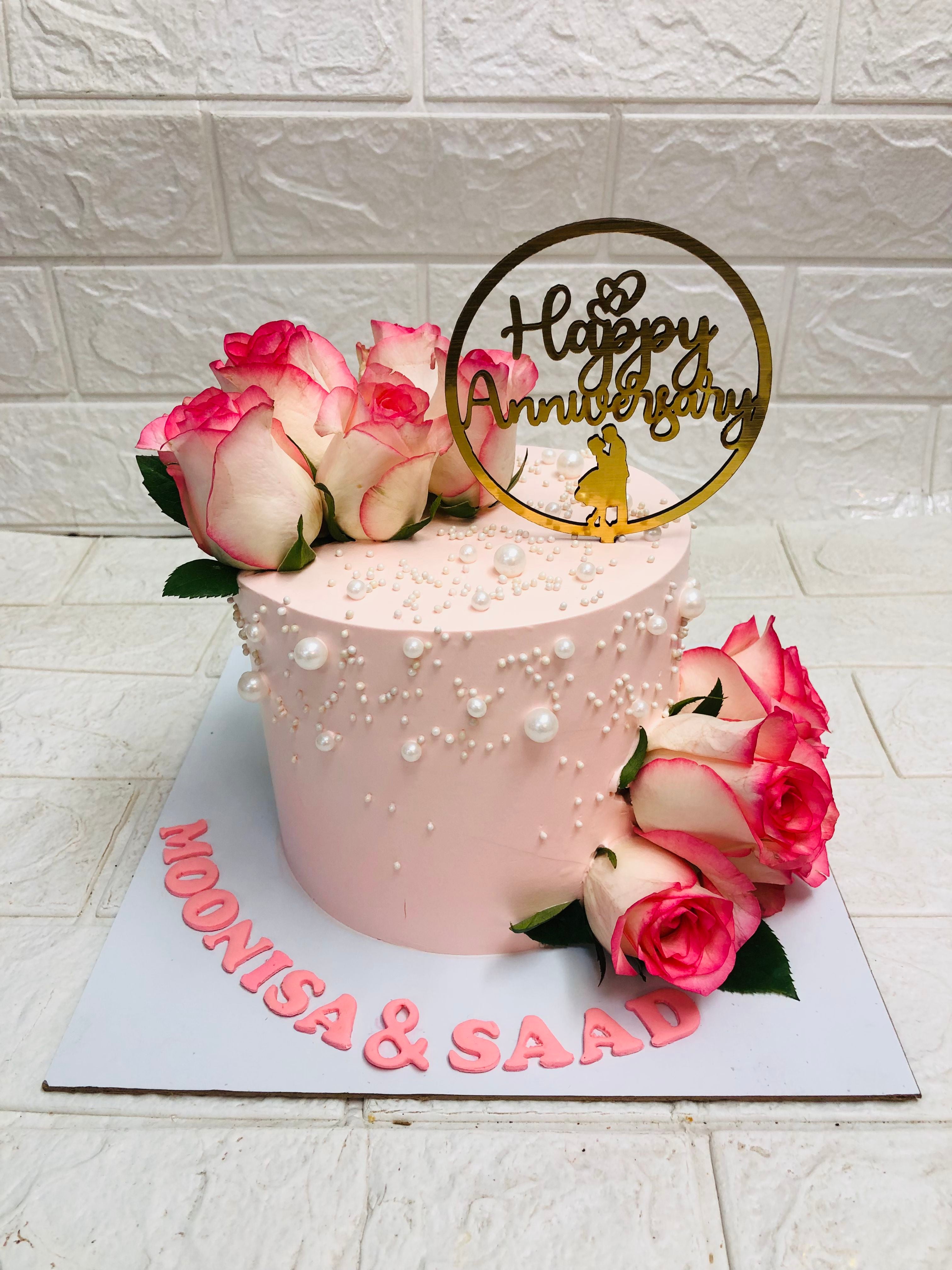 Elegant pink anniversary cake with smooth frosting, white pearl-like decorations, and fresh roses with white petals and pink edges. The cake features a golden topper reading 'Happy Anniversary' with a couple's silhouette and the names 'MOONISA & SAAD' written in pink icing on a white cake board.