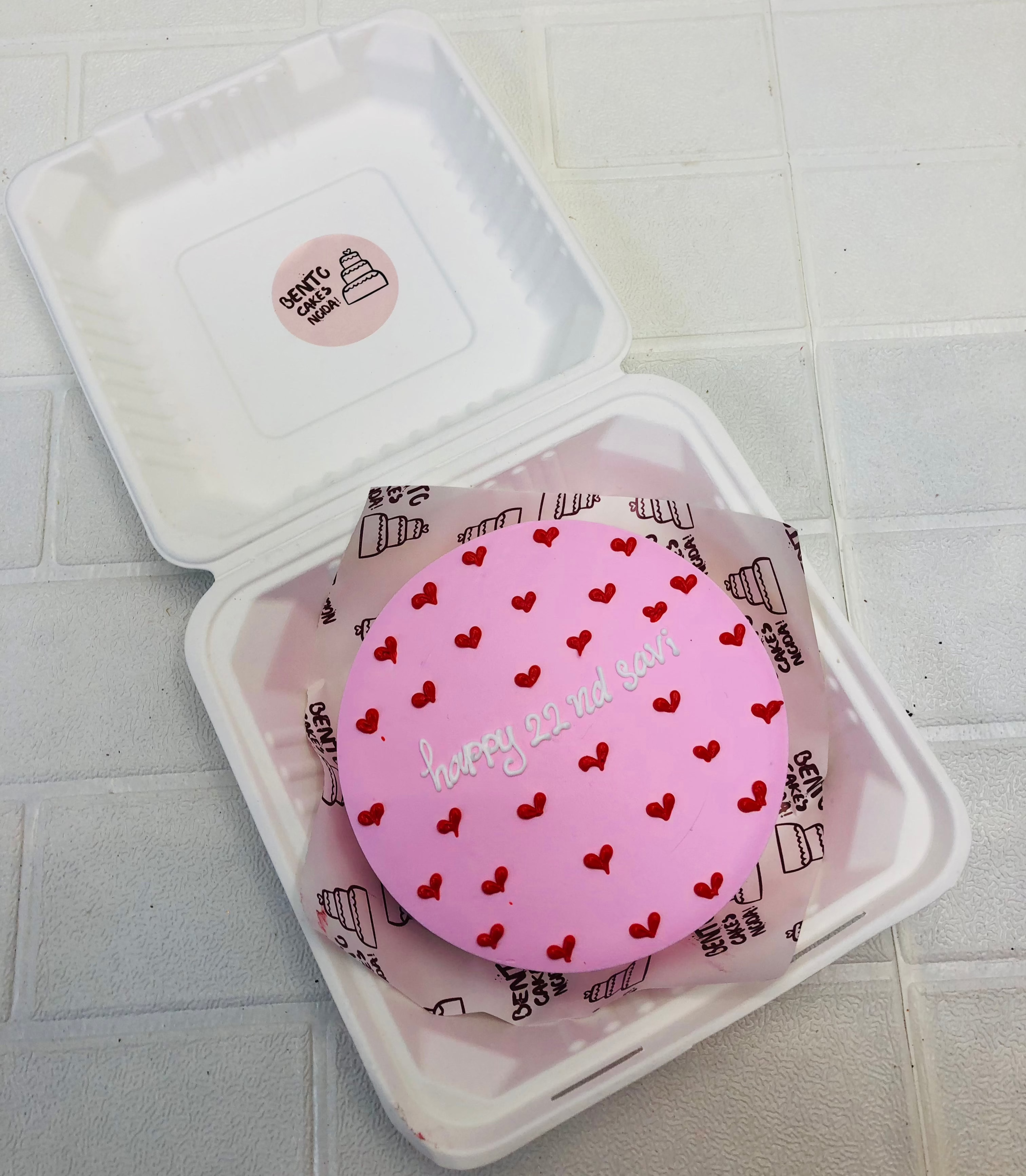 A pink bento cake having red hearts design all over the cake and a text "Happy 22 nd savi" written with white icing.