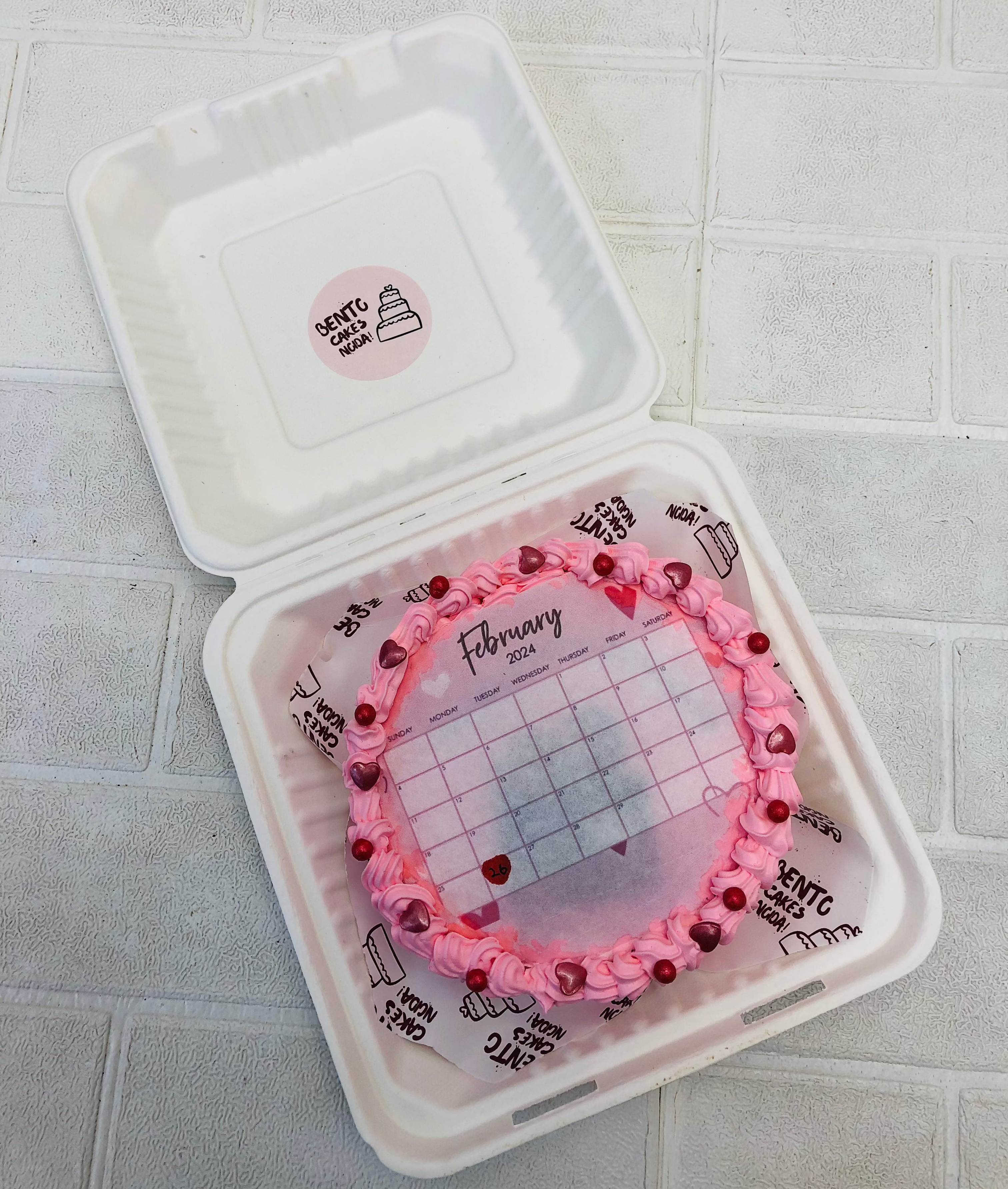 A light pink round bento cake with february calendar theme and also decorated with red hearts and red sprinkles.