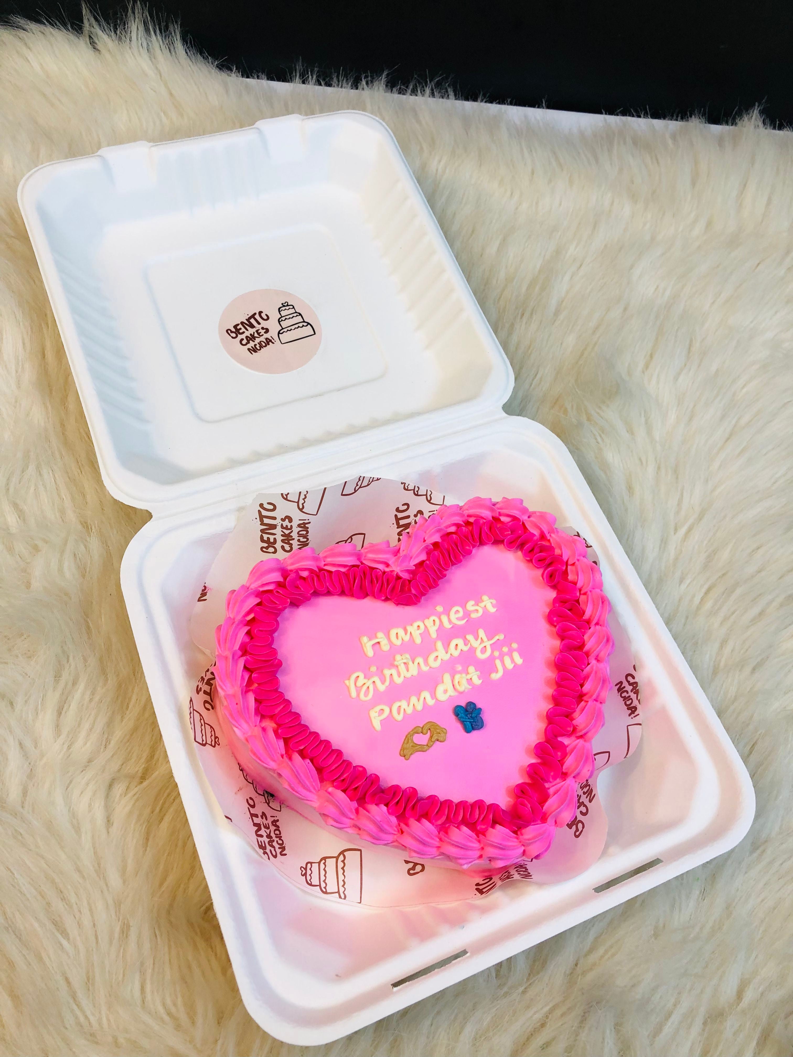 A light pink designer heart shape cake with dark pink design on its ounday and a "Happy Birthday Pandit jii" written with offwhite color and cute emojies made on it.