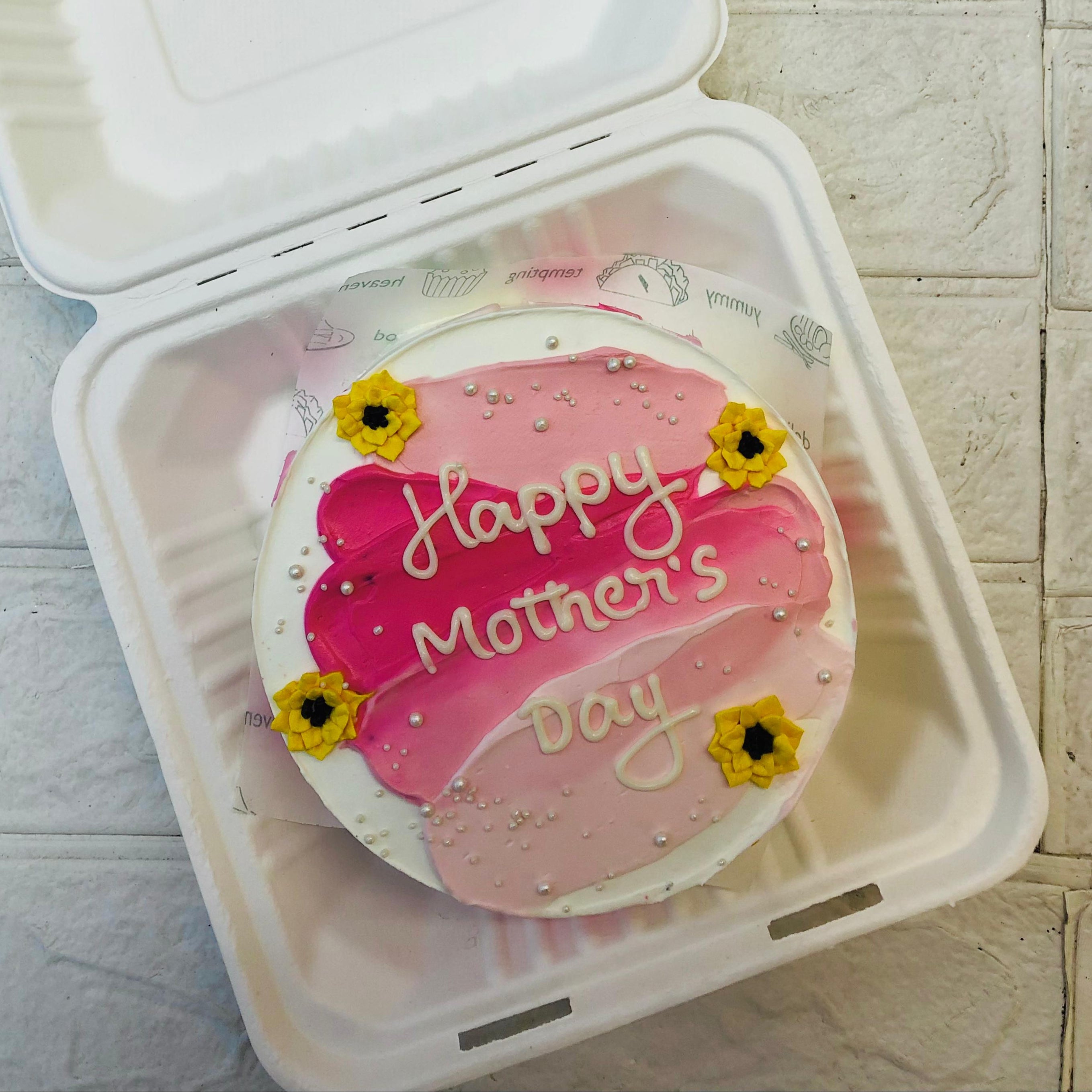 A pink shades round cake with yellow florals design on it and 'Happy Mother's Day" Text written with whitw icing.