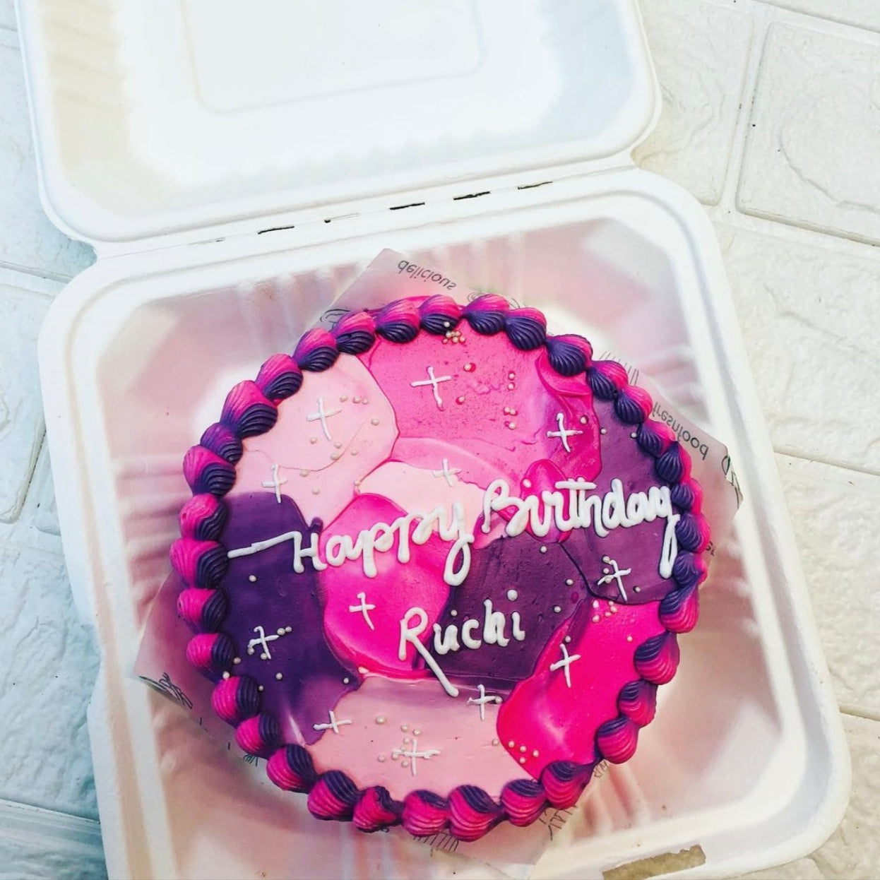 A beautifully decorated birthday cake with a pink and purple gradient design, placed inside a white takeout box. The cake has the message "Happy Birthday Ruchi" written in white icing.