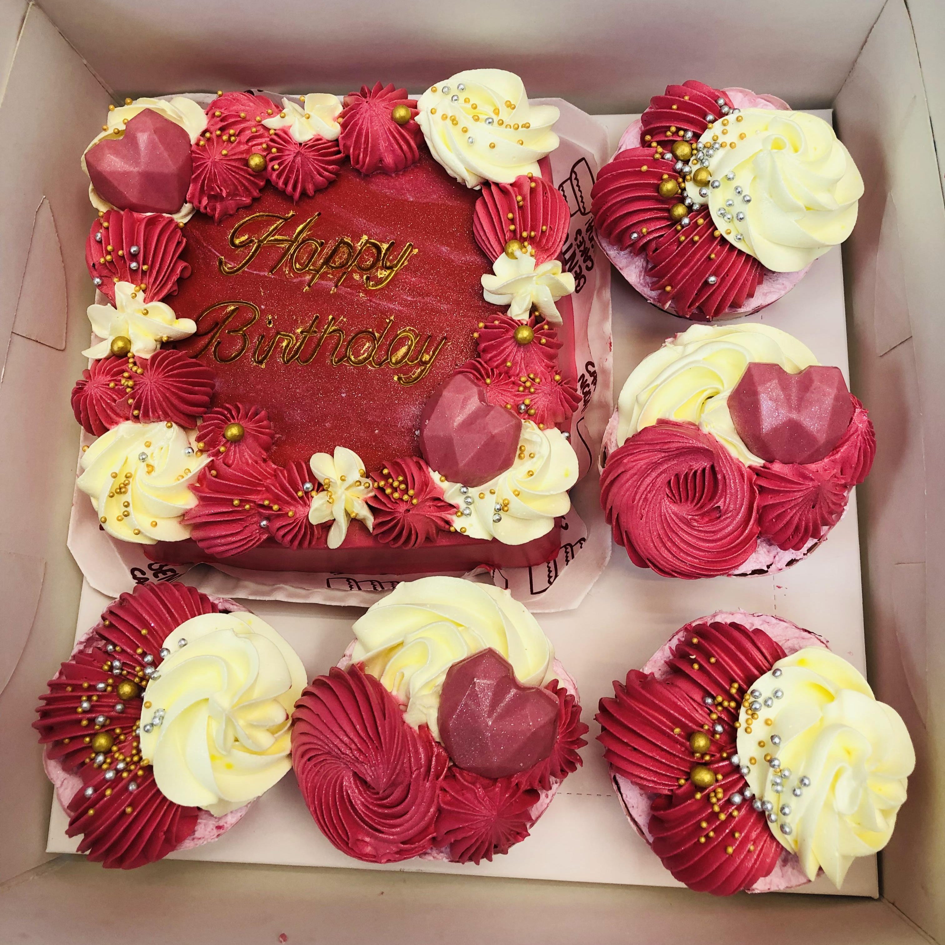 A square birthday cake with red and white frosting, adorned with gold beads and floral accents, accompanied by cupcakes featuring matching red and white frosting and heart-shaped decorations.