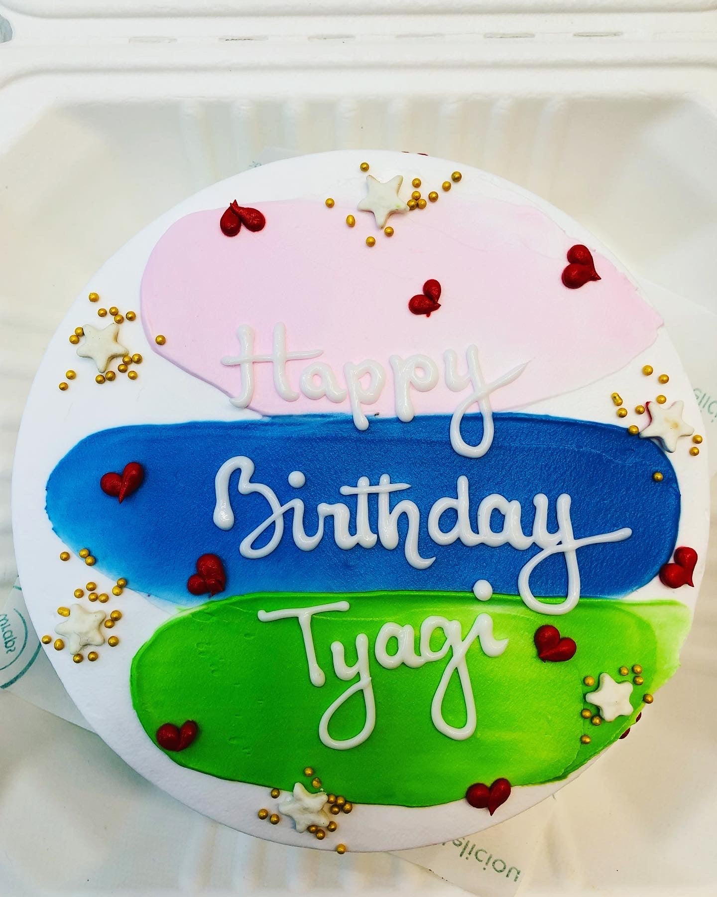 A white cake with blue, green and light pink icing on it and some red hearts with golden sprinkle beautifully decorated on it.