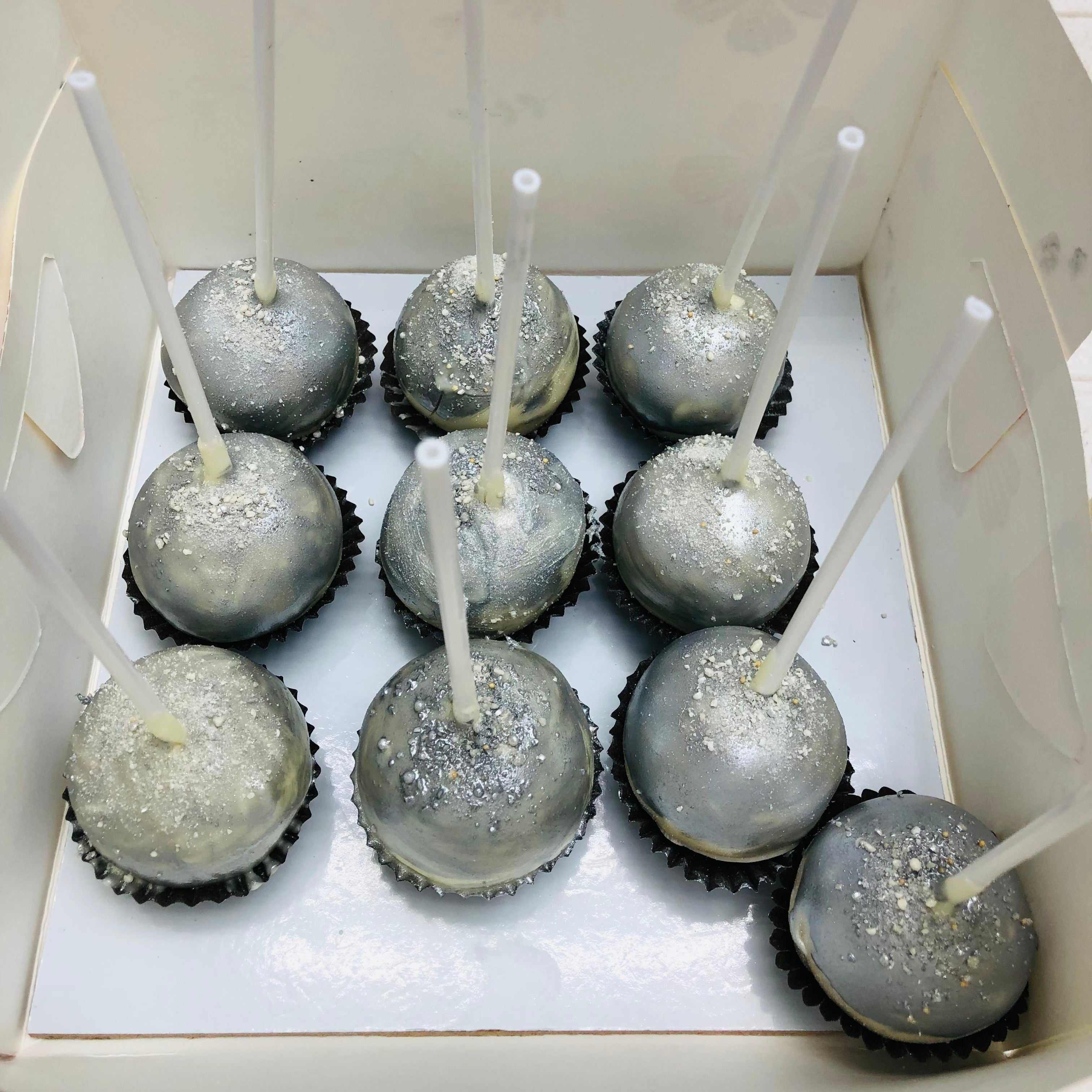 A box containing nine silver-colored cake pops with white sticks, arranged in three rows of three. Each cake pop is decorated with a shimmering, metallic finish and sprinkled with edible glitter.