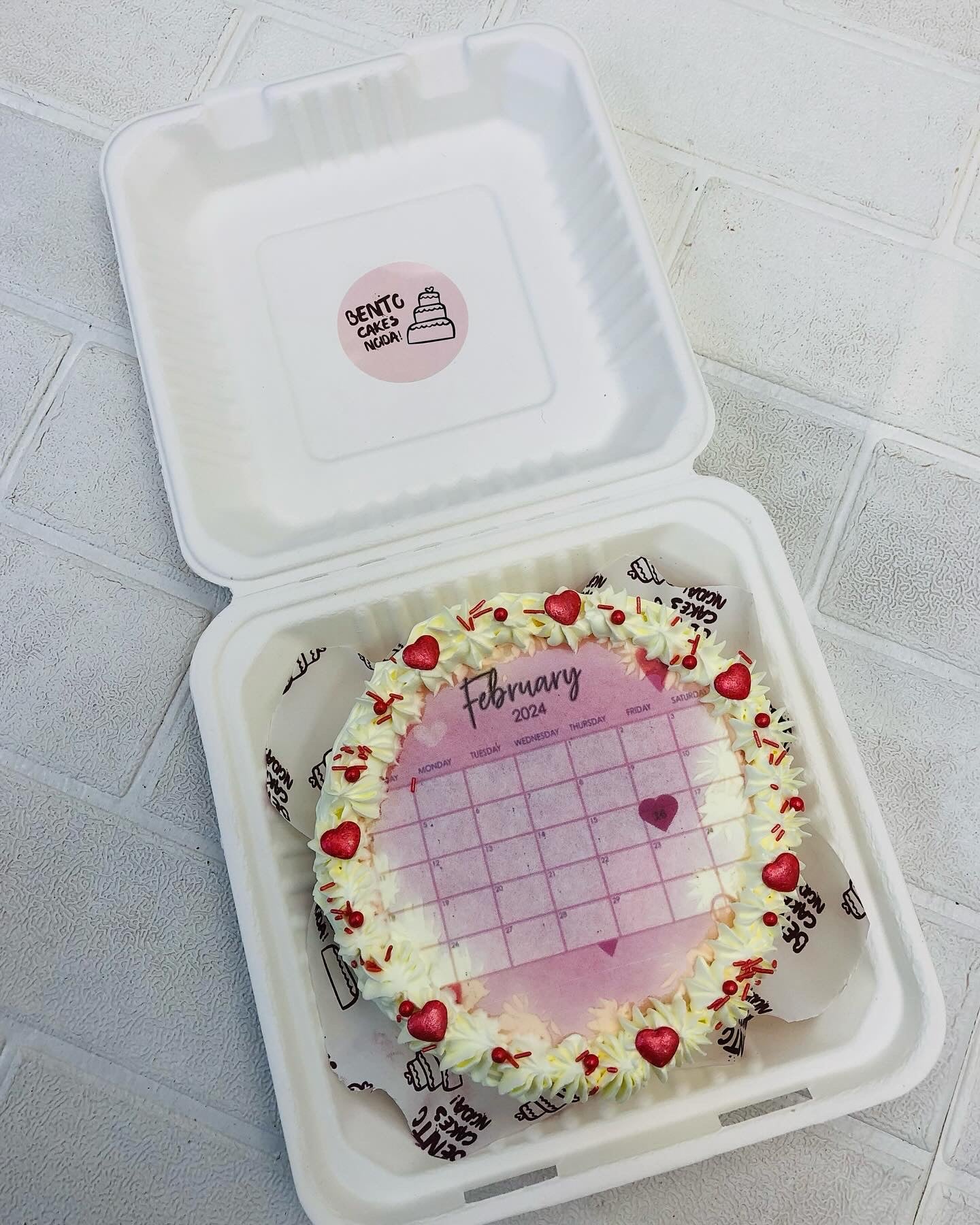 An off white fire bento cake with february fire calendar and its border decorated with red hearts and sprinkles.