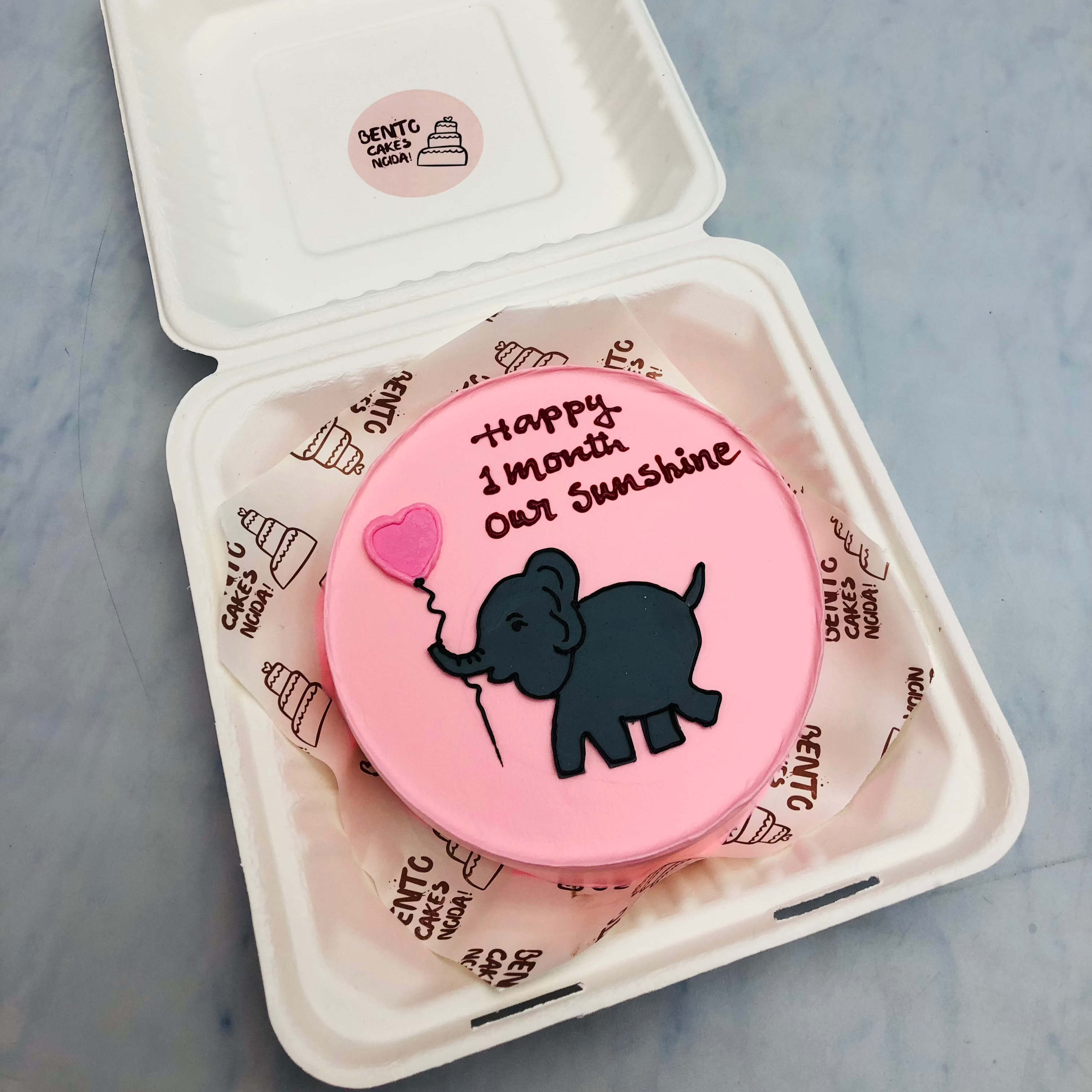 A light pink bento cake with a black baby elefant drawing takken a pink balloon for a baby girl birthday.