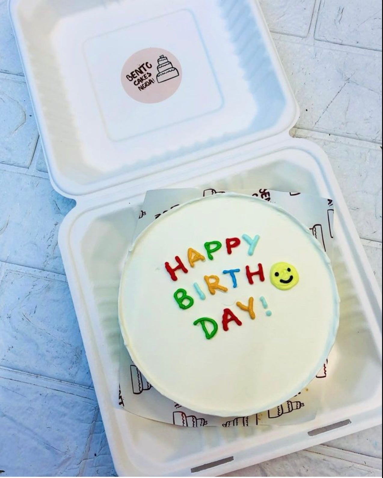 A white cake in a bento box descoated with colorful text of "HAPPY BIRTHDAY!" anda smily emoji on it.