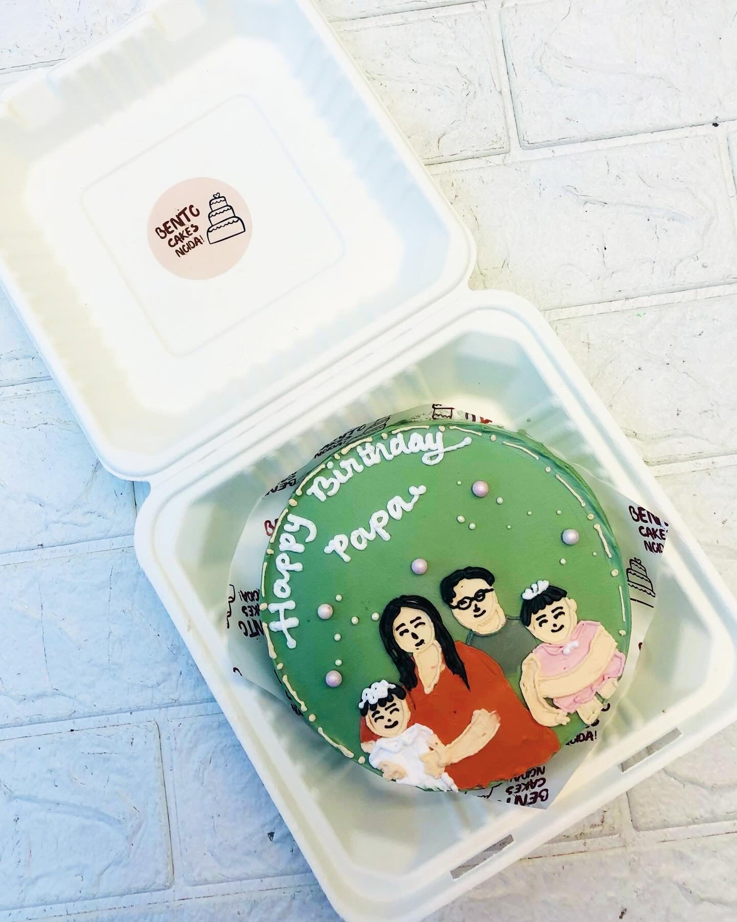 A custom-made birthday cake with green icing, featuring an illustration of a family. The cake is placed inside a white bento box and has 'Happy Birthday Papa' written on it, making it a unique and heartfelt gift."
