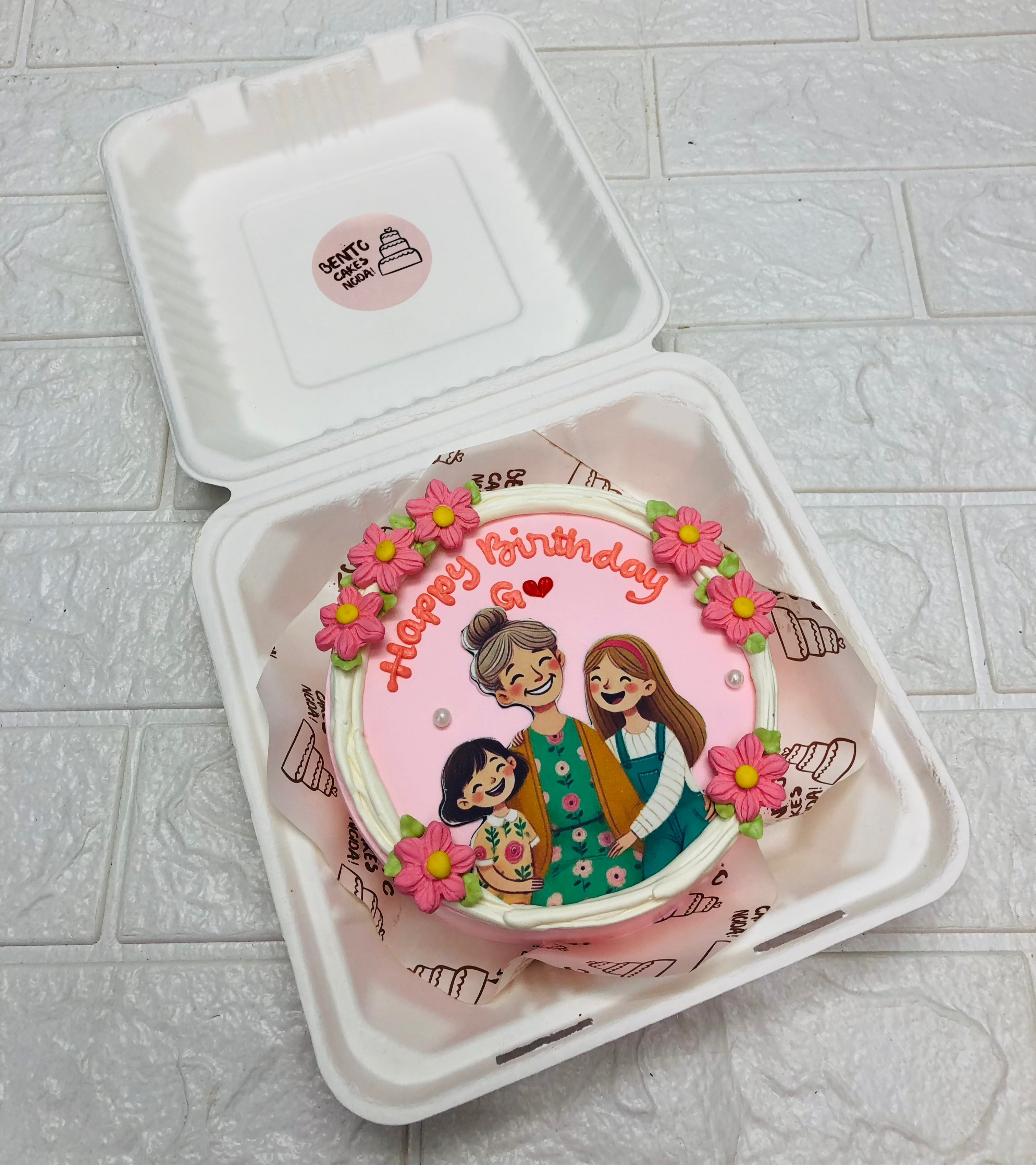 A birthday cake featuring an illustration of three people, decorated with pink flowers and the words 'Happy Birthday G' in red icing.