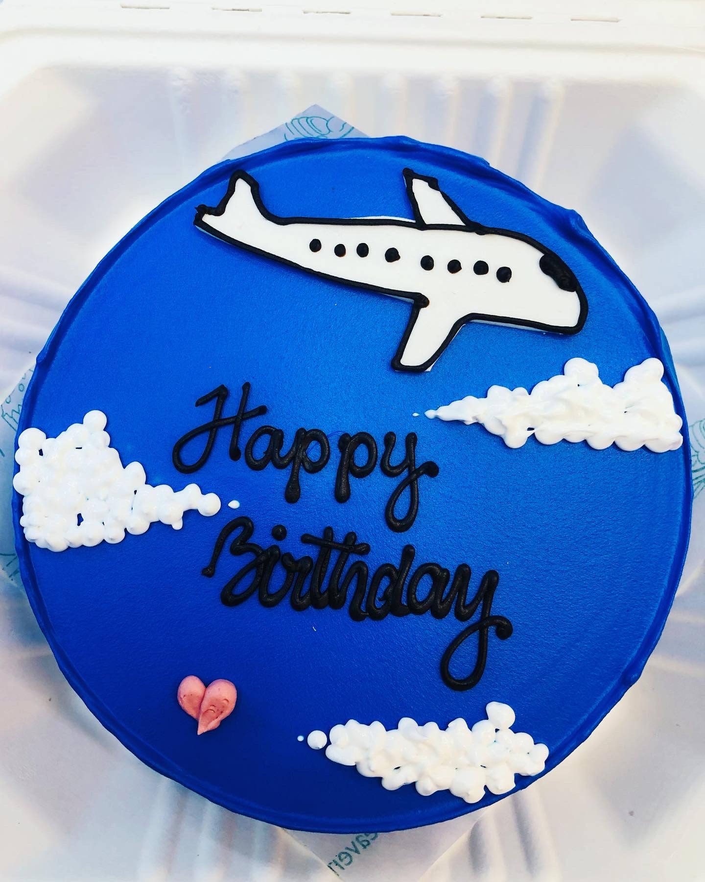 A round blue birthday cake with a white airplane and clouds decoration, and the words "Happy Birthday" written in black icing.