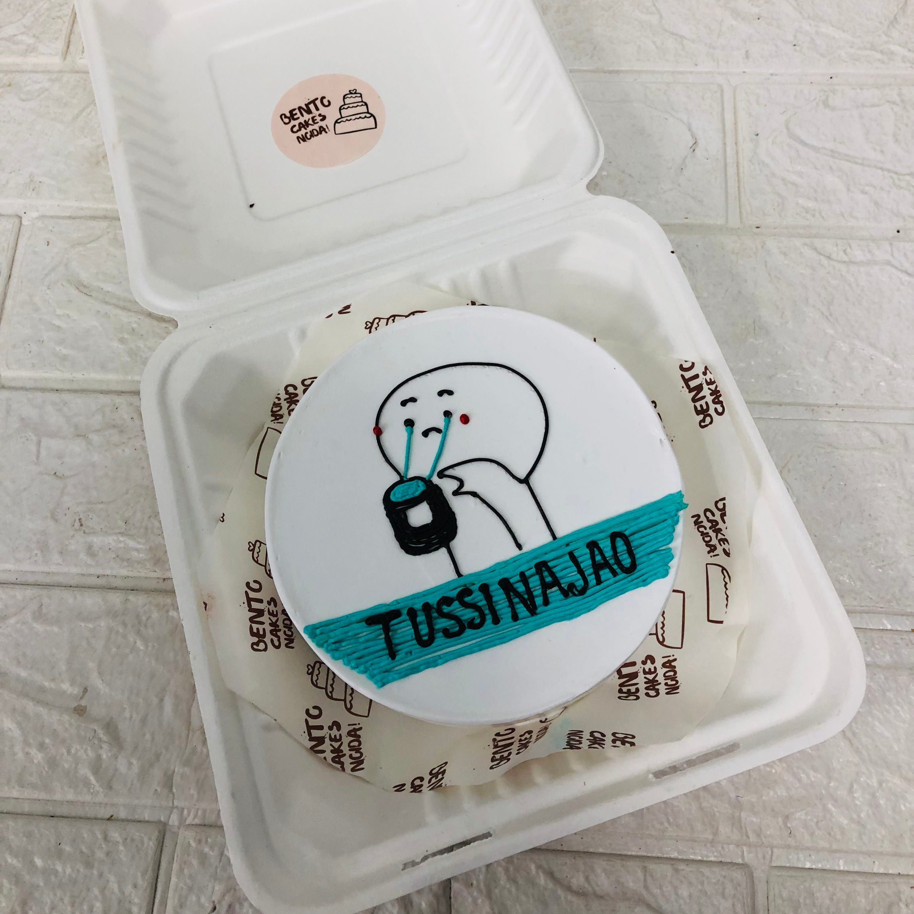 A white cake in a bento box with a weeping cartoon character saying "TUSSI NA JAO" 