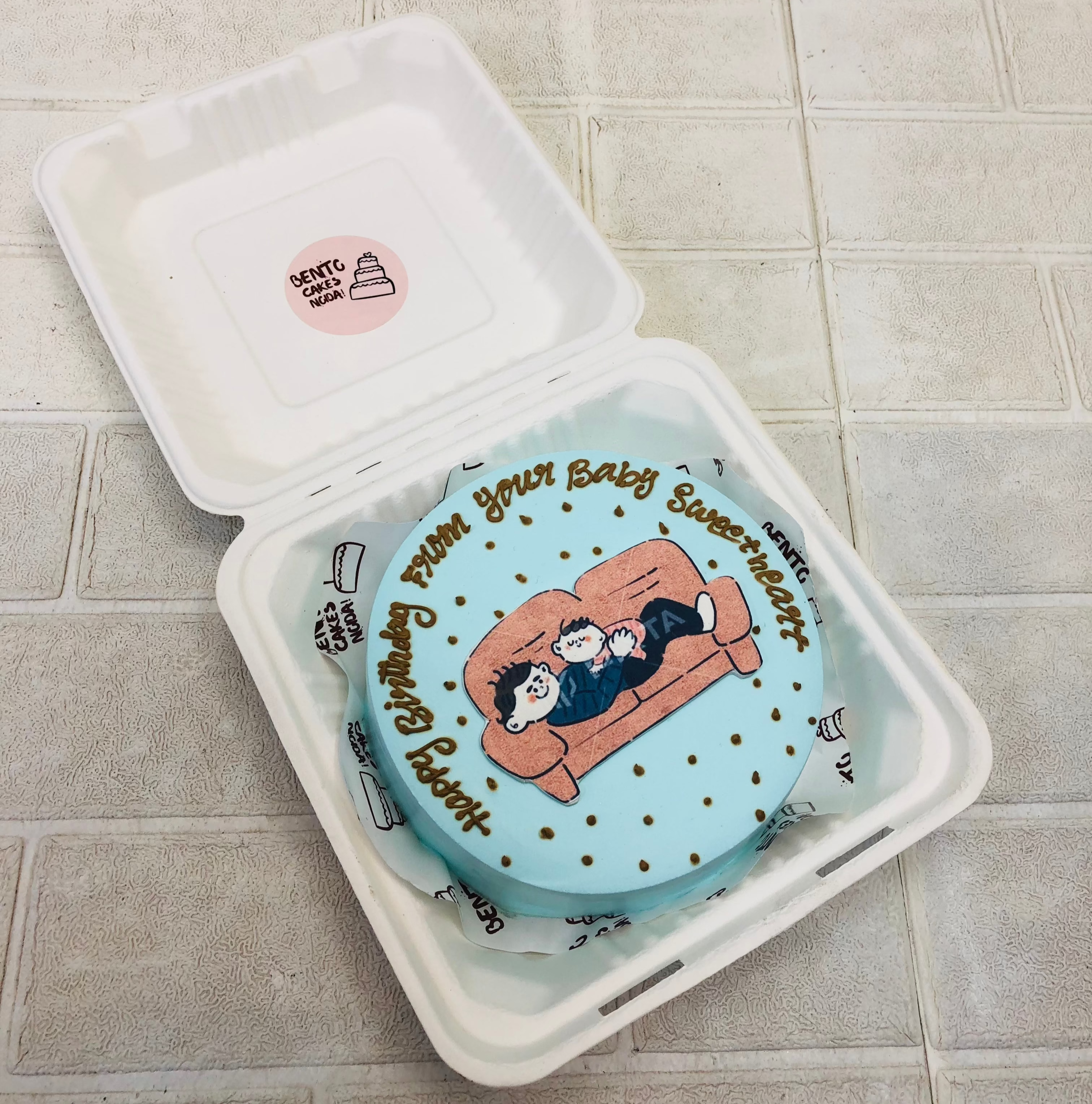A cool blue bento cake is designed with a father and baby laying on sofa print placed on cake with a Birthday wishing text.