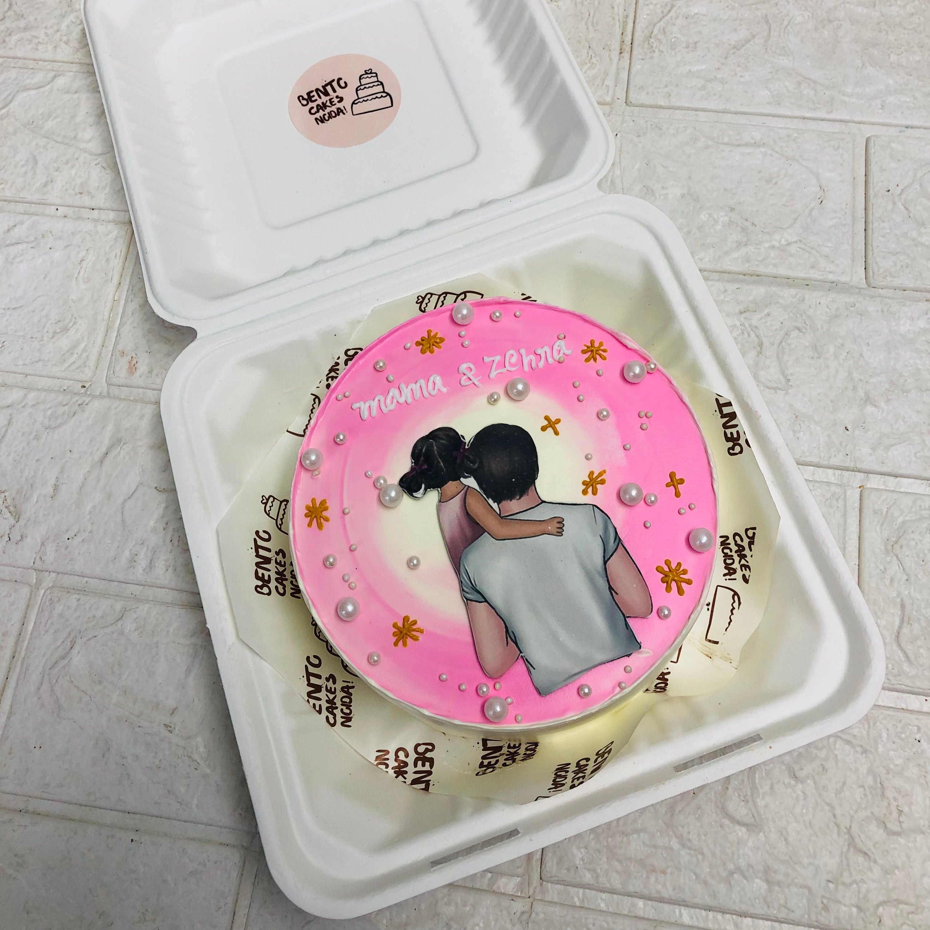 A round cake with pink frosting, decorated with an illustration of a person carrying a child, and the text "mama & Zehra" on top.