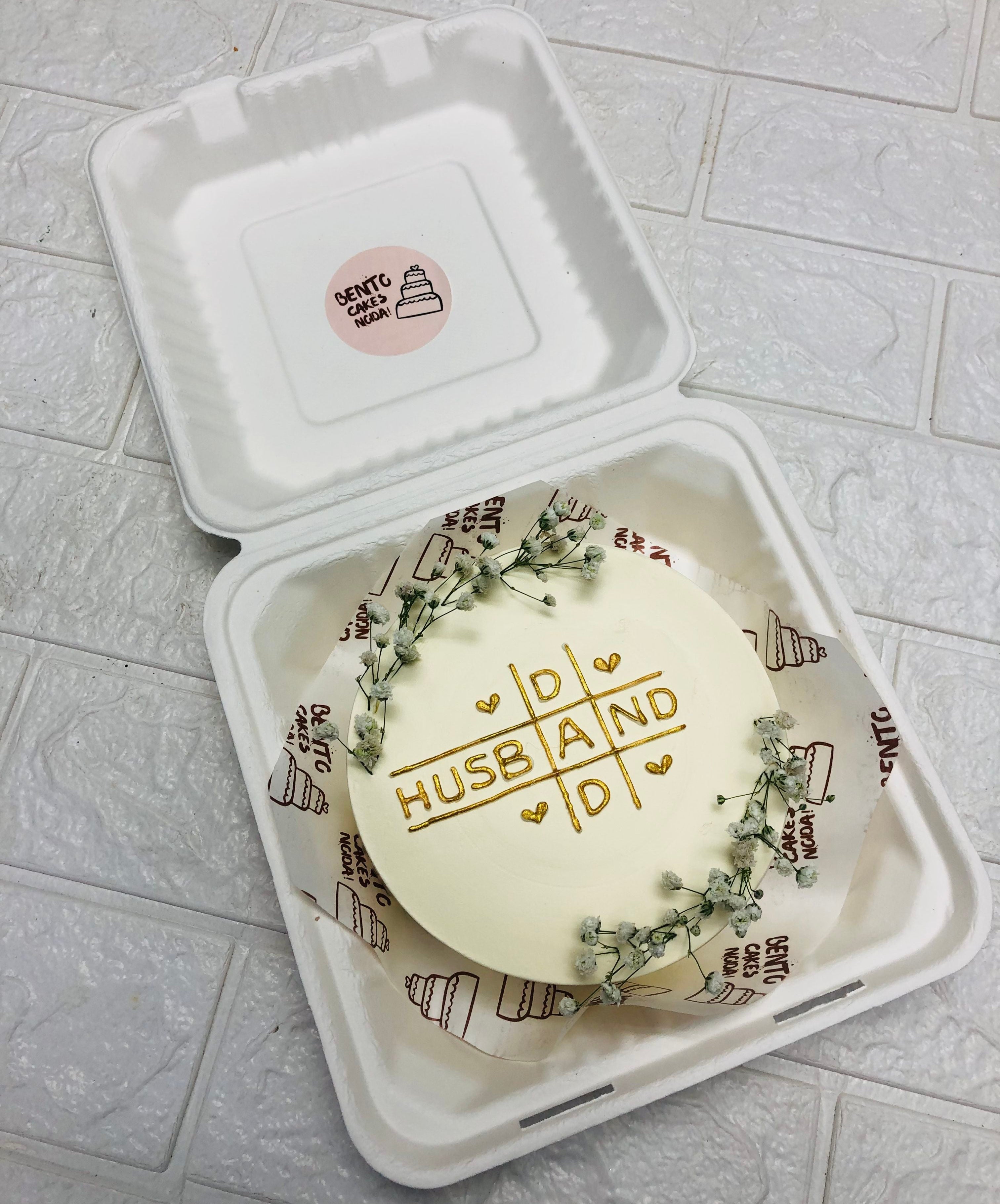 A beautifully decorated bento cake, featuring a tic-tac-toe grid design with the words "HUSBAND" and "DAD" written in gold icing, surrounded by small hearts. The cake is encircled by delicate white flowers for an elegant touch.