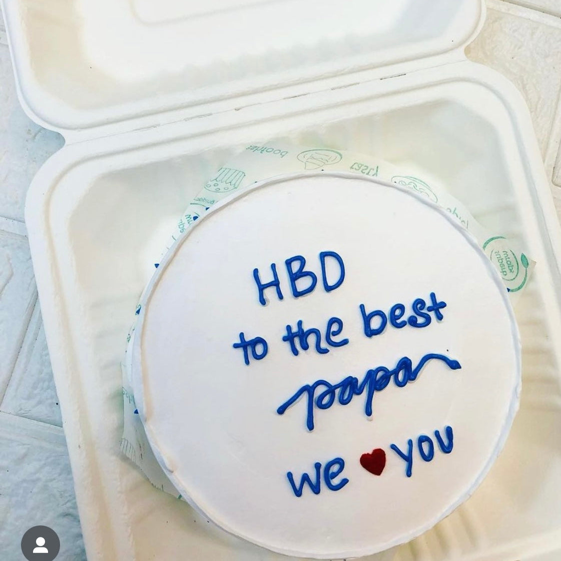A white cake with blue icing that reads "HBD to the best papa we ❤️ you" inside a white  bento box.