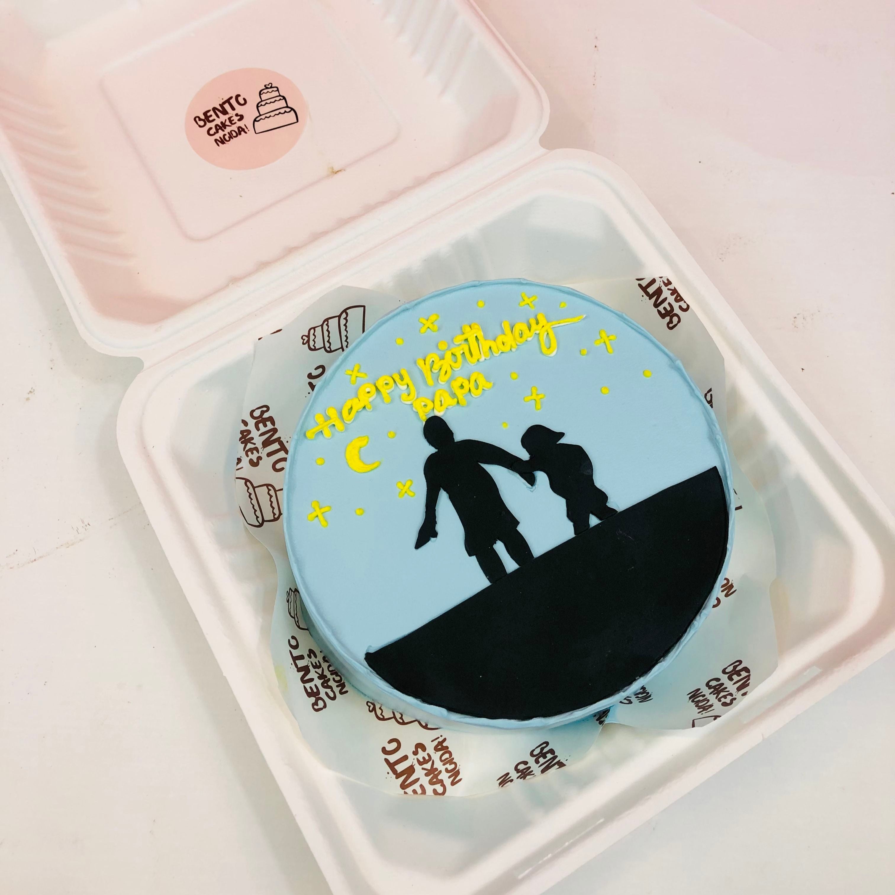 A round light blue birthday cake inside a white Bento Cakes box, featuring a black silhouette of an adult and a child holding hands on a hill, surrounded by yellow stars and a crescent moon, with "Happy Birthday Papa" written in yellow icing.