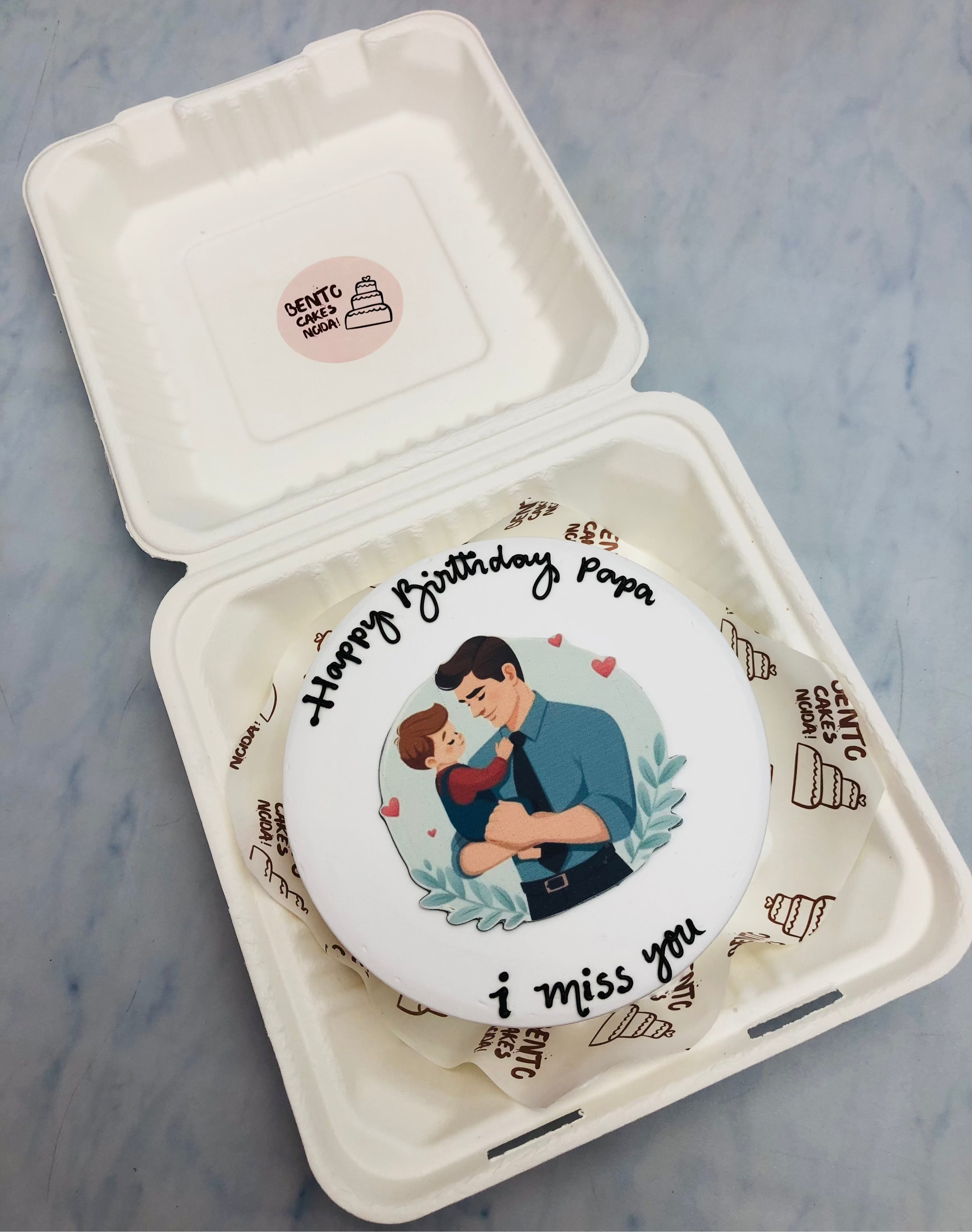 A round cake with a touching illustration of a father embracing his child, surrounded by delicate hearts and leaves. The cake is enclosed in a white box with the heartfelt messages 'Happy Birthday Papa' and 'I miss you' written on it.