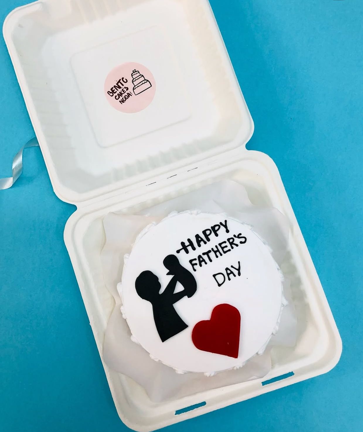 A Father's Day cake in a white biodegradable container. The cake has a white fondant base with a black silhouette of a father holding a child, the text "HAPPY FATHER'S DAY" in black, and a red heart. 