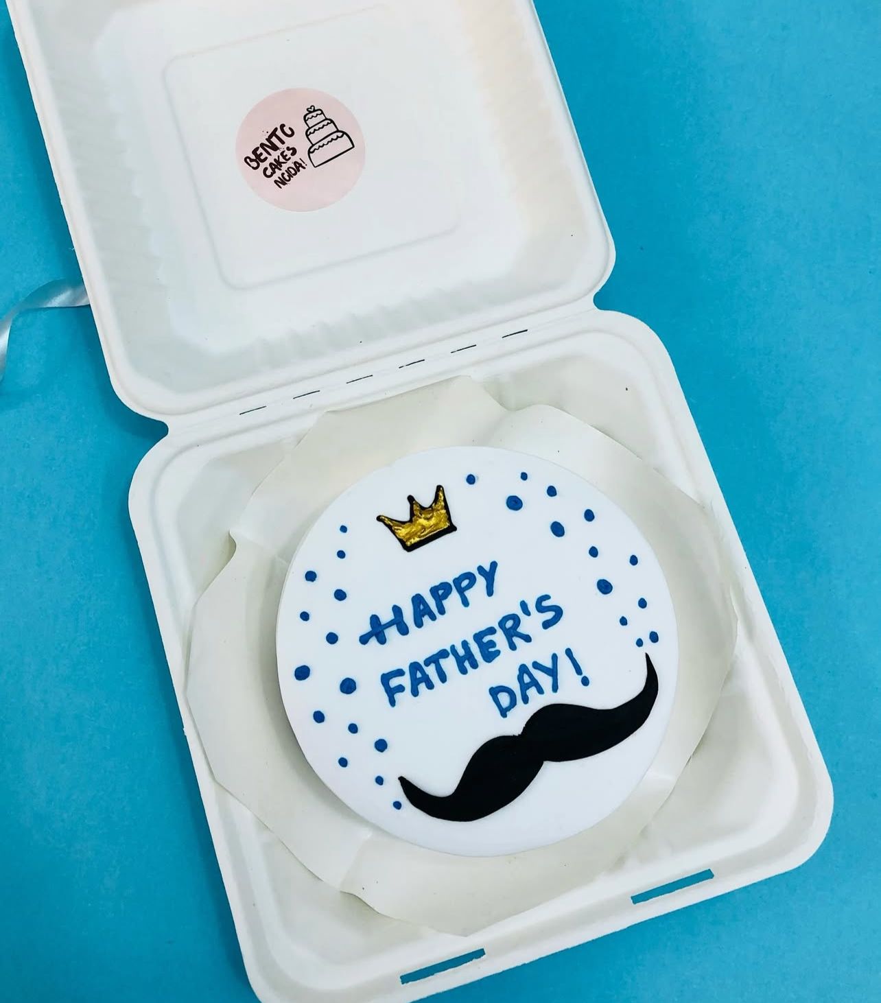 A round cookie decorated with a golden crown, a black mustache, and the message "HAPPY FATHER'S DAY!" in blue icing, surrounded by blue dots. 
