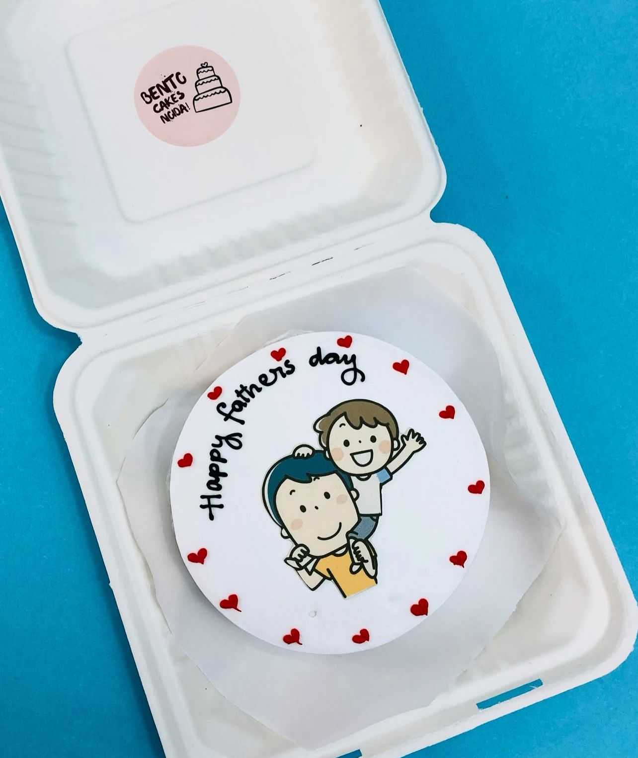 A Father's Day cake from Bentocakes Ngopi, featuring an illustration of a father carrying a child on his shoulders, surrounded by small red hearts and the text "Happy father's day" in a playful handwritten style. 