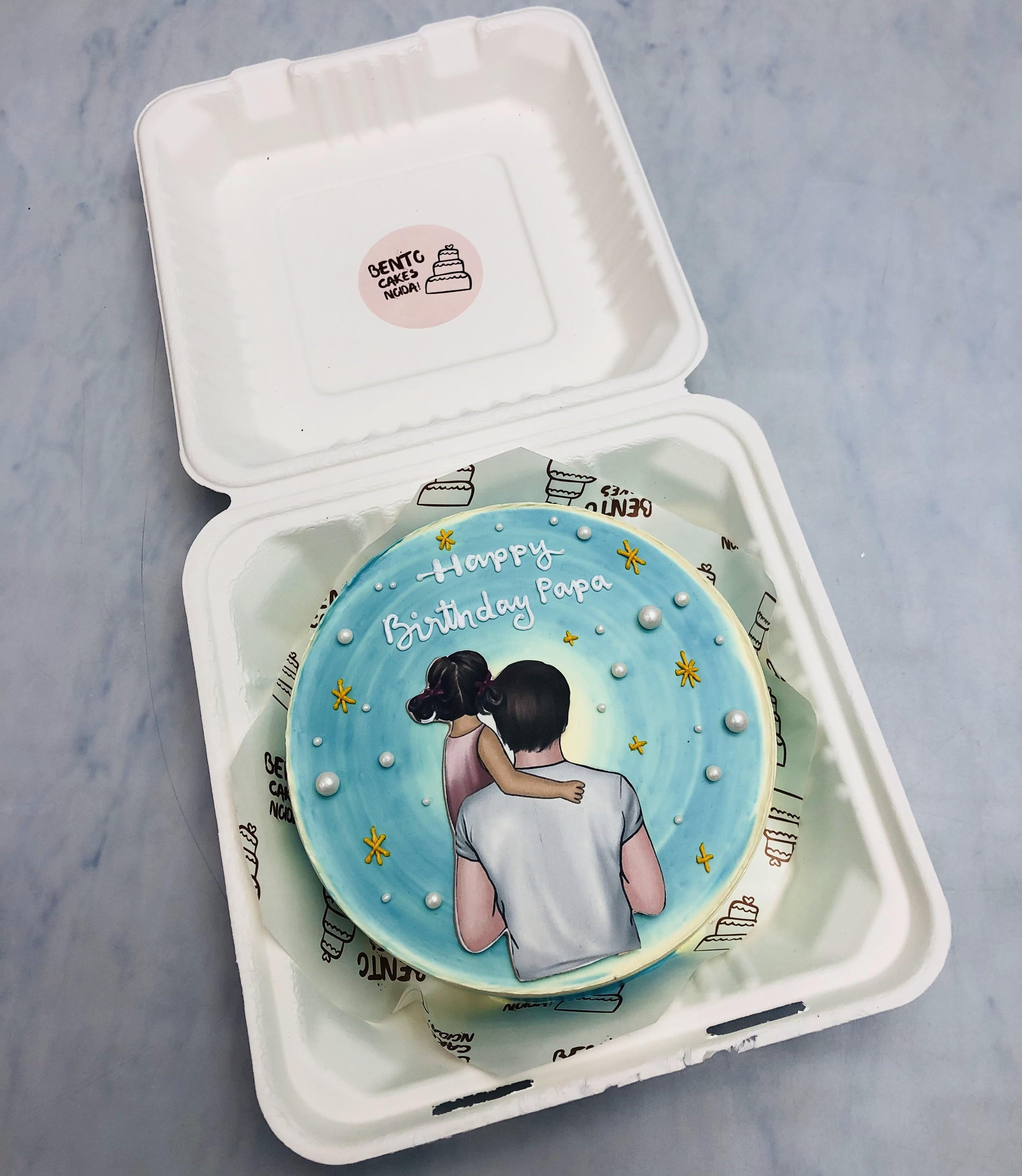 An ombre blue design on white bento cake decorated with white sprinkles with a girl and father print on it.