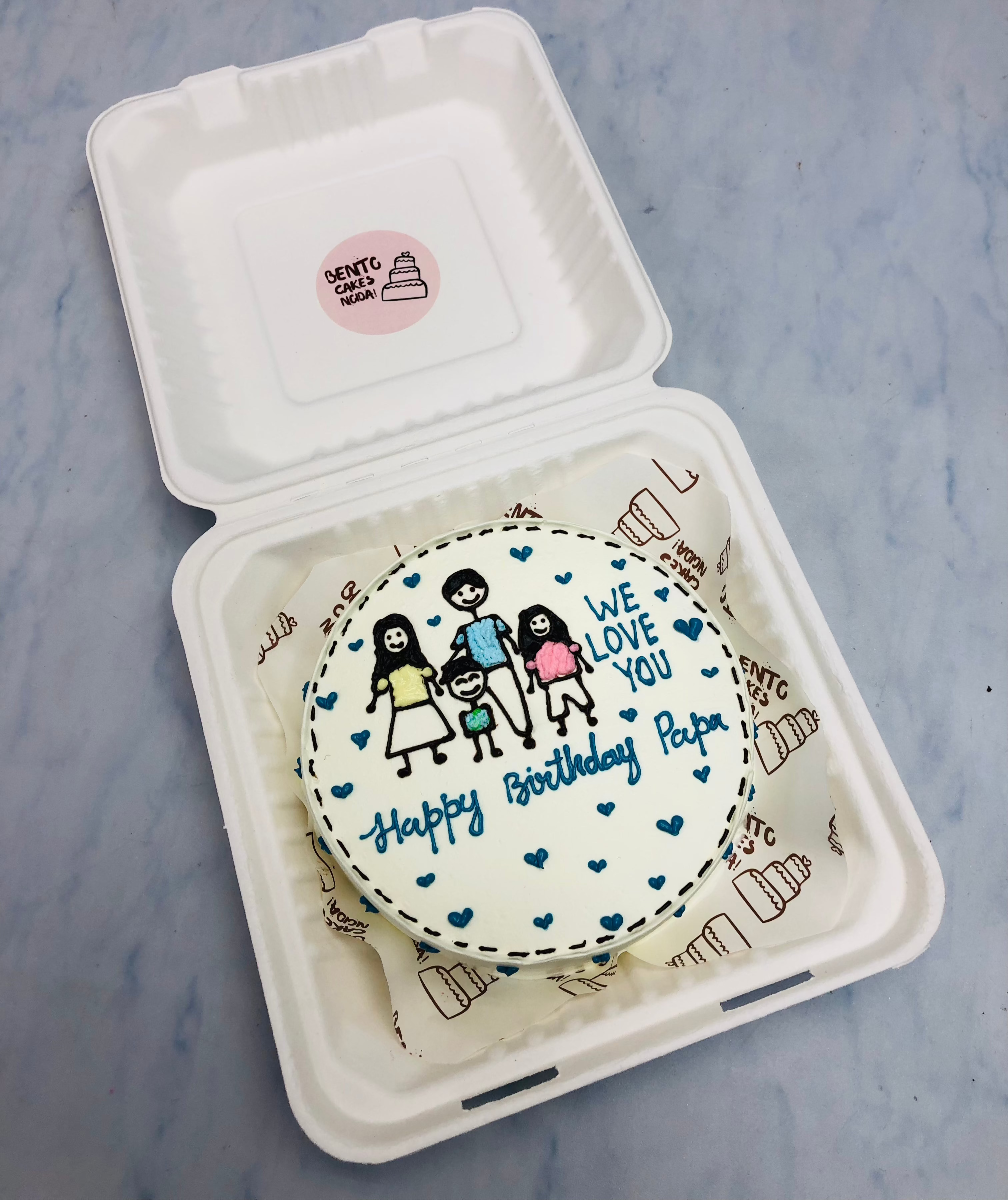 A round birthday cake with white icing, featuring a hand-drawn illustration of a family of four. The cake is placed inside a white bento box and decorated with blue hearts. The message “WE LOVE YOU” is written above the family, while “Happy Birthday Papa” is below.
